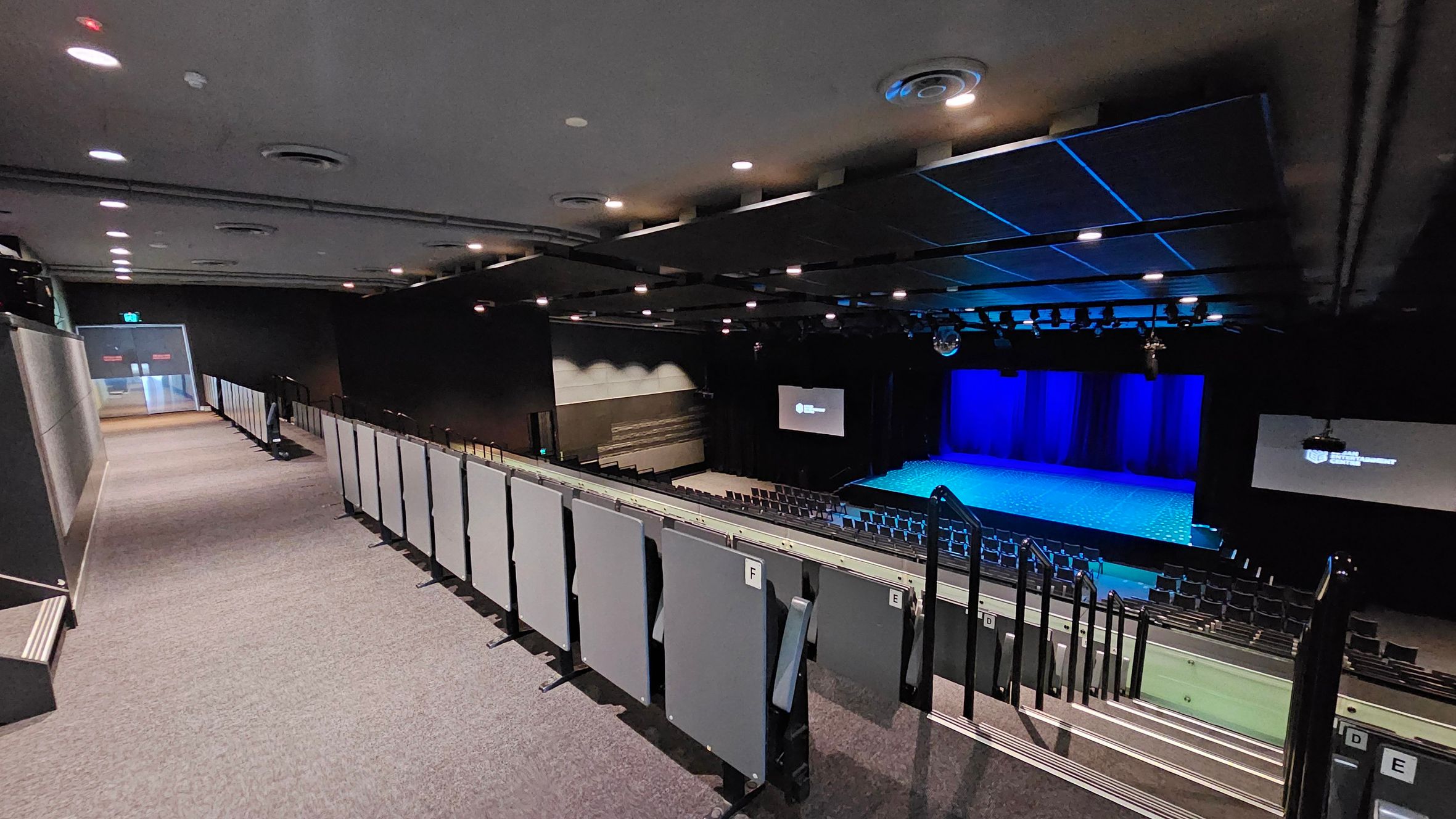A theatre interior with a view from the upper seating area. On the left, a carpeted walkway leads to an exit door. The right side features tiered seating with grey panels and handrails. In the middle, a stage with a blue-lit backdrop is visible. Two screens flank the stage, displaying text. The ceiling is equipped with recessed lighting and ventilation ducts. The floor is carpeted, and the overall atmosphere is dimly lit. A theatre interior with a view from the upper seating area. On the left, a carpeted walkway leads to an exit door. The right side features tiered seating with grey panels and handrails. In the middle, a stage with a blue-lit backdrop is visible. Two screens flank the stage, displaying text. The ceiling is equipped with recessed lighting and ventilation ducts. The floor is carpeted, and the overall atmosphere is dimly lit.
