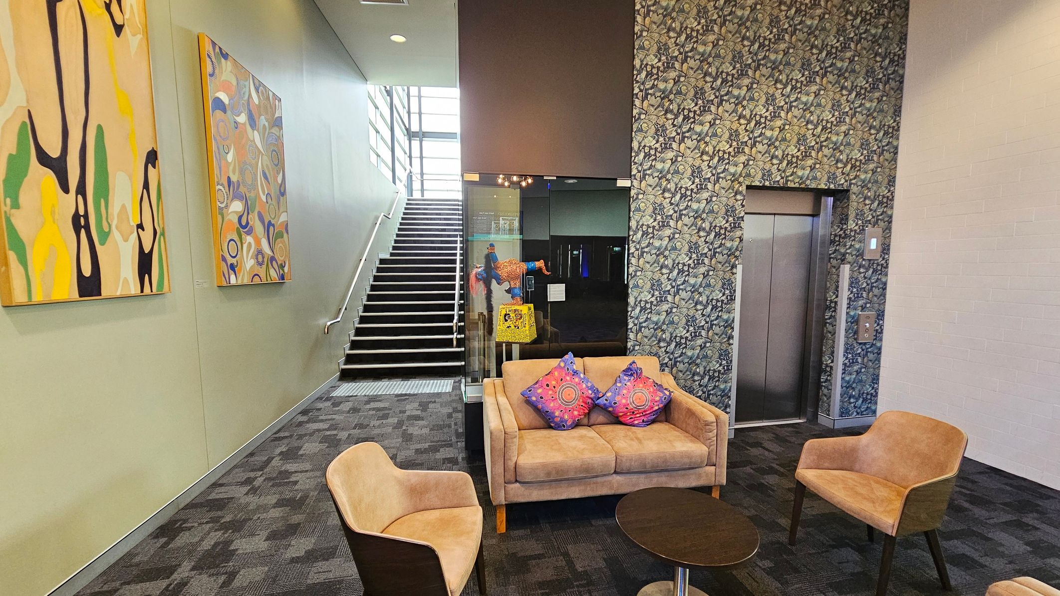 A modern interior space with a staircase on the left, featuring two abstract paintings on the wall. In the centre, a glass partition displays a colourful artwork. A seating area includes a beige sofa with two patterned cushions and two matching chairs around a small round table. On the right, an elevator is set into a wall with a floral pattern. The floor is carpeted with a dark geometric design. A modern interior space with a staircase on the left, featuring two abstract paintings on the wall. In the centre, a glass partition displays a colourful artwork. A seating area includes a beige sofa with two patterned cushions and two matching chairs around a small round table. On the right, an elevator is set into a wall with a floral pattern. The floor is carpeted with a dark geometric design.