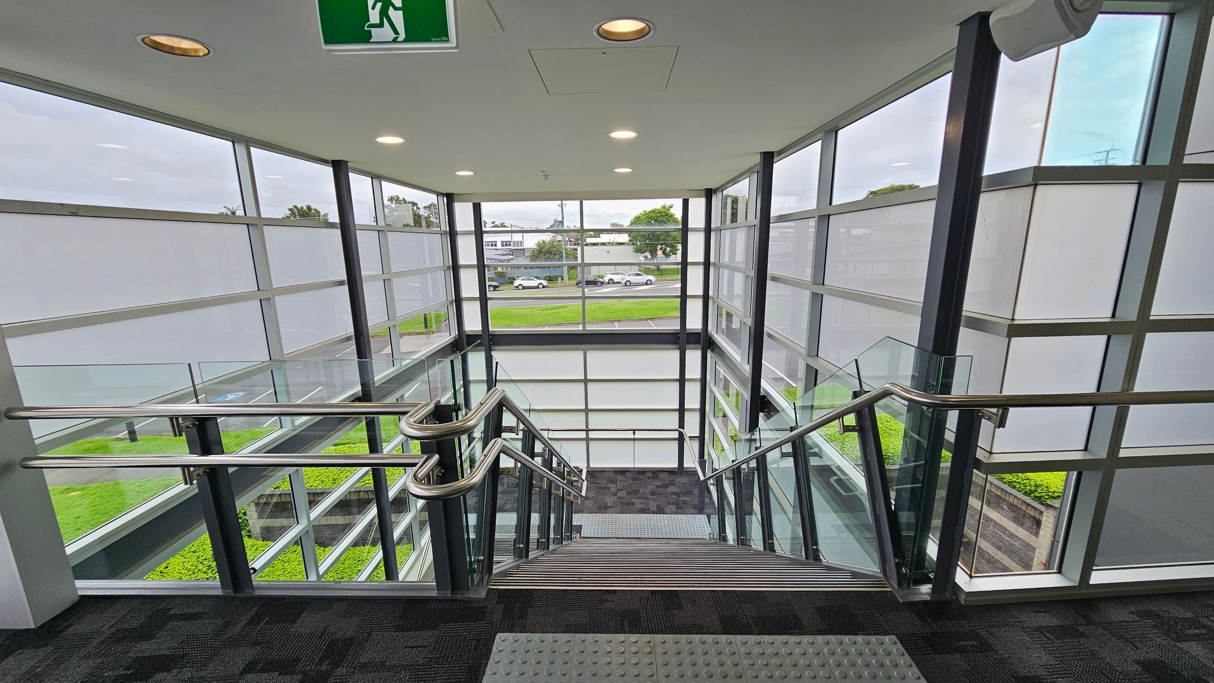 A modern indoor staircase with metal railings and glass panels on both sides. The walls are made of large glass windows, providing a view of the outside, including a road with parked cars and greenery. The ceiling is white with recessed lighting and an emergency exit sign. The floor is carpeted in a dark pattern, and there are tactile paving tiles at the top of the stairs. A modern indoor staircase with metal railings and glass panels on both sides. The walls are made of large glass windows, providing a view of the outside, including a road with parked cars and greenery. The ceiling is white with recessed lighting and an emergency exit sign. The floor is carpeted in a dark pattern, and there are tactile paving tiles at the top of the stairs.