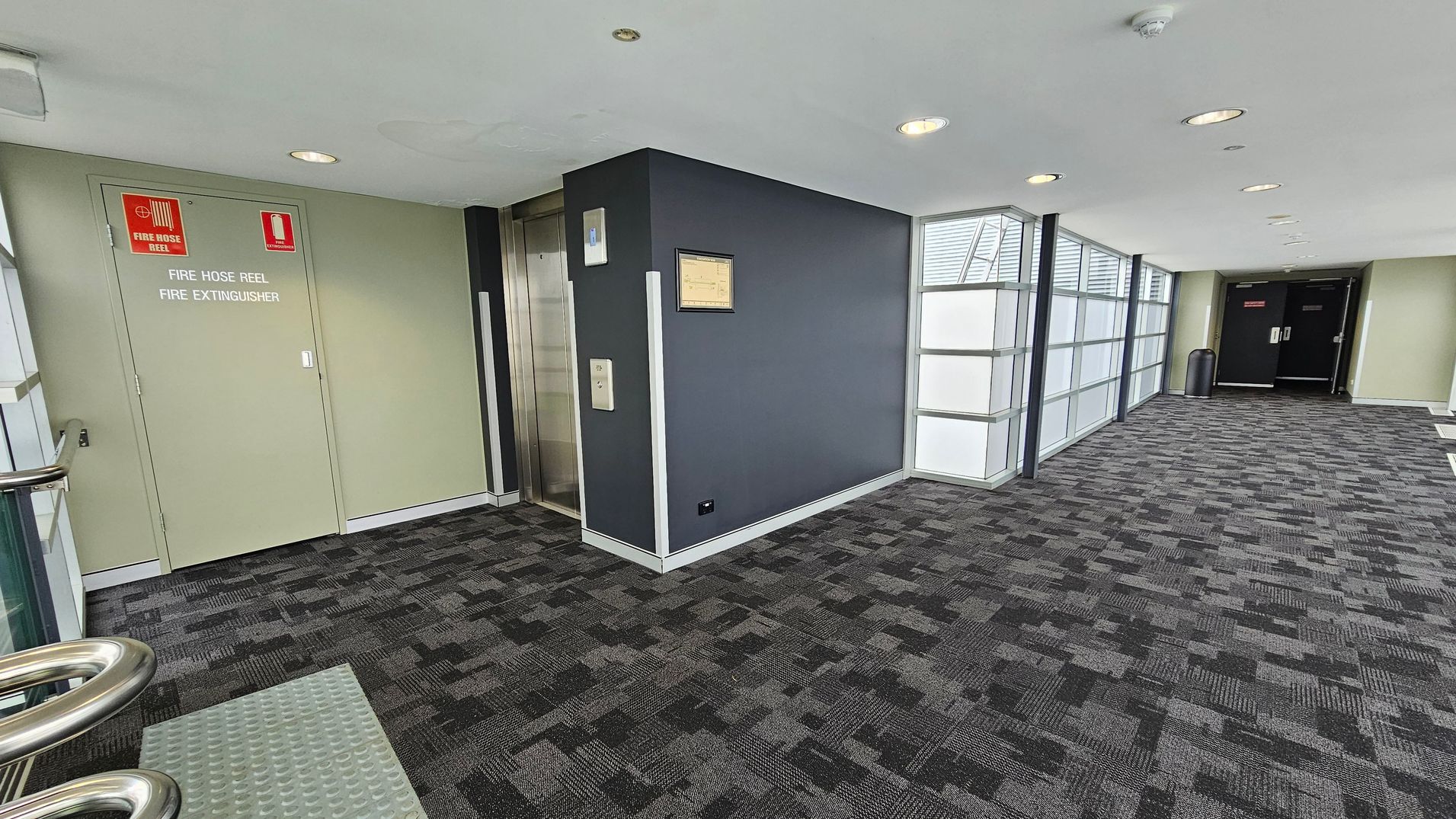 A corridor with a patterned grey carpet. On the left, there is a green door marked for a fire hose reel and extinguisher. Next to it is a lift with a metallic door. The wall beside the lift is dark grey with a framed notice. On the right, large windows with frosted glass panels allow light in. At the end of the corridor, there are double black doors with a bin nearby. The ceiling is white with recessed lights. A corridor with a patterned grey carpet. On the left, there is a green door marked for a fire hose reel and extinguisher. Next to it is a lift with a metallic door. The wall beside the lift is dark grey with a framed notice. On the right, large windows with frosted glass panels allow light in. At the end of the corridor, there are double black doors with a bin nearby. The ceiling is white with recessed lights.
