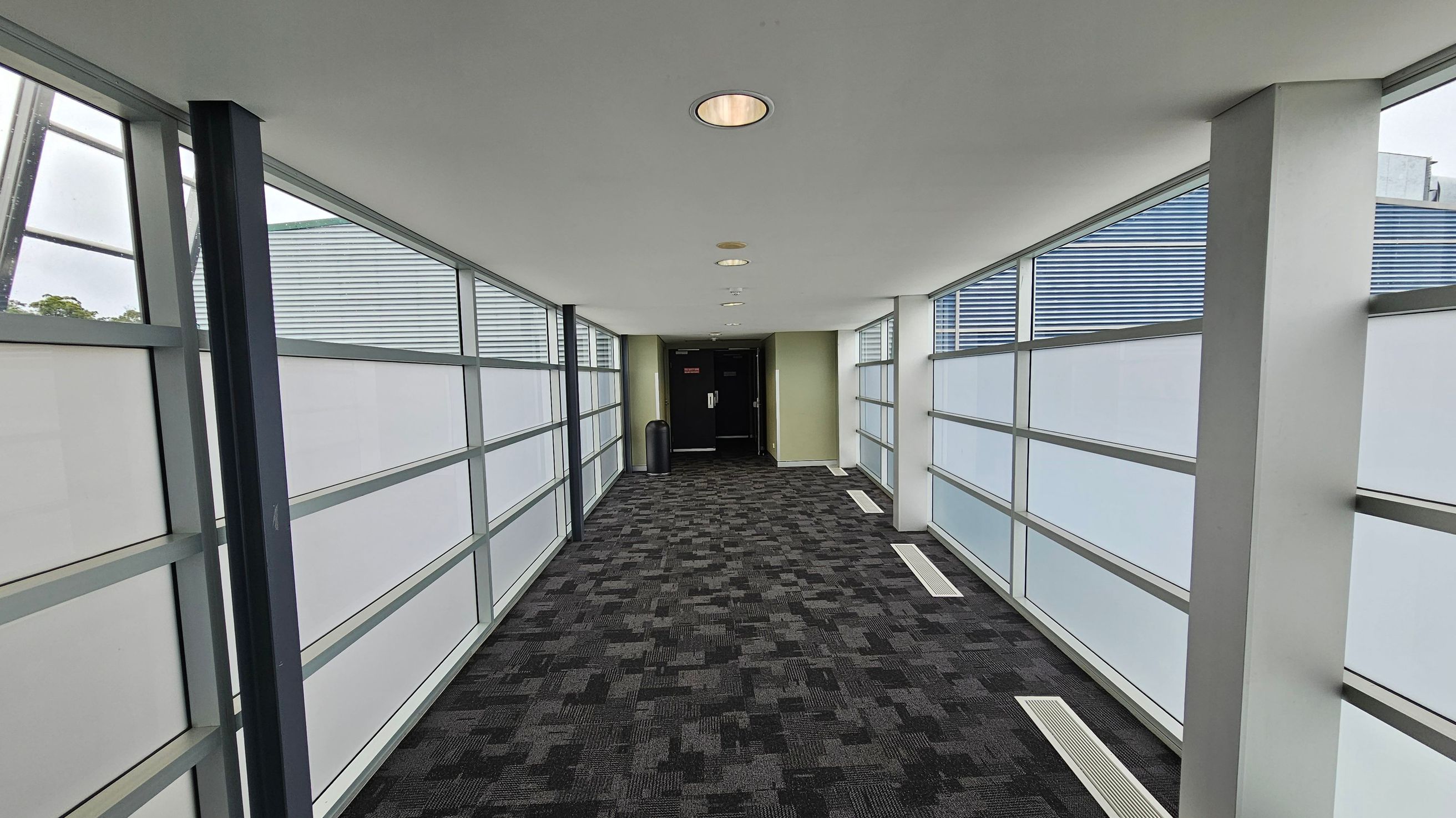 A corridor with large frosted glass windows on both sides, framed by metal supports. The floor is covered with a dark patterned carpet. The ceiling is white with recessed circular lights. At the end of the corridor, there is a black door and a green wall, with a black bin nearby. A corridor with large frosted glass windows on both sides, framed by metal supports. The floor is covered with a dark patterned carpet. The ceiling is white with recessed circular lights. At the end of the corridor, there is a black door and a green wall, with a black bin nearby.