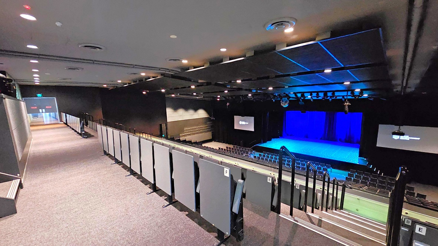 A theatre interior with a view from the upper seating area. On the left, a carpeted walkway leads to an exit door. The right side features tiered seating with grey panels and handrails. In the middle, a stage with a blue-lit backdrop is visible, flanked by two large screens. The ceiling is equipped with recessed lighting and ventilation ducts. The floor is carpeted, and the overall atmosphere is dimly lit. A theatre interior with a view from the upper seating area. On the left, a carpeted walkway leads to an exit door. The right side features tiered seating with grey panels and handrails. In the middle, a stage with a blue-lit backdrop is visible, flanked by two large screens. The ceiling is equipped with recessed lighting and ventilation ducts. The floor is carpeted, and the overall atmosphere is dimly lit.