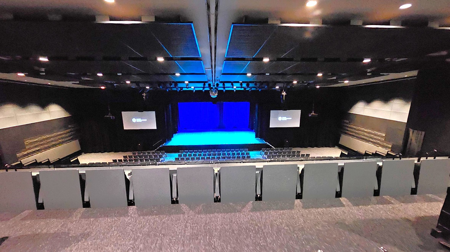 A large auditorium with tiered seating in the foreground, leading down to a stage with a blue-lit backdrop. Two screens are positioned on either side of the stage, displaying a logo. The walls are dark with acoustic panels, and the ceiling is equipped with numerous recessed lights. The floor is carpeted in a dark colour, matching the overall dim and professional atmosphere of the space. A large auditorium with tiered seating in the foreground, leading down to a stage with a blue-lit backdrop. Two screens are positioned on either side of the stage, displaying a logo. The walls are dark with acoustic panels, and the ceiling is equipped with numerous recessed lights. The floor is carpeted in a dark colour, matching the overall dim and professional atmosphere of the space.