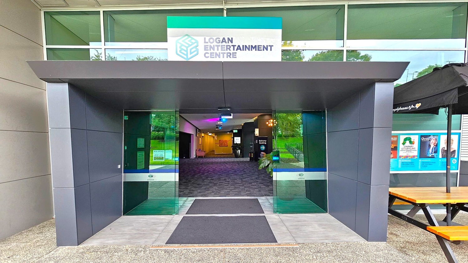A modern entrance to the Logan Entertainment Centre with a grey and green colour scheme. The entrance features large glass doors with a sign above displaying the centre's name and logo. Inside, a carpeted hallway is visible, leading to various rooms. To the right, there is a wooden picnic table and a display of posters under a black canopy. The exterior walls are clad in grey panels, and the ground is covered with a textured surface. A modern entrance to the Logan Entertainment Centre with a grey and green colour scheme. The entrance features large glass doors with a sign above displaying the centre's name and logo. Inside, a carpeted hallway is visible, leading to various rooms. To the right, there is a wooden picnic table and a display of posters under a black canopy. The exterior walls are clad in grey panels, and the ground is covered with a textured surface.