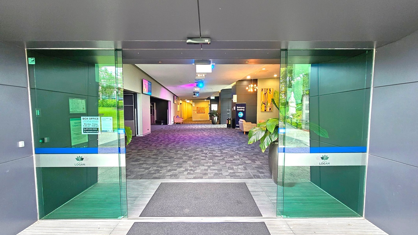 Glass doors with a blue and white stripe, labelled "Logan," open into a spacious lobby. The floor is covered with a patterned carpet. On the right, a potted plant and a sign for "Balcony Seating" are visible. The left side has a box office window with opening hours displayed. The ceiling features recessed lighting and colourful spotlights. In the background, there are more seating areas and artwork on the walls. Glass doors with a blue and white stripe, labelled "Logan," open into a spacious lobby. The floor is covered with a patterned carpet. On the right, a potted plant and a sign for "Balcony Seating" are visible. The left side has a box office window with opening hours displayed. The ceiling features recessed lighting and colourful spotlights. In the background, there are more seating areas and artwork on the walls.