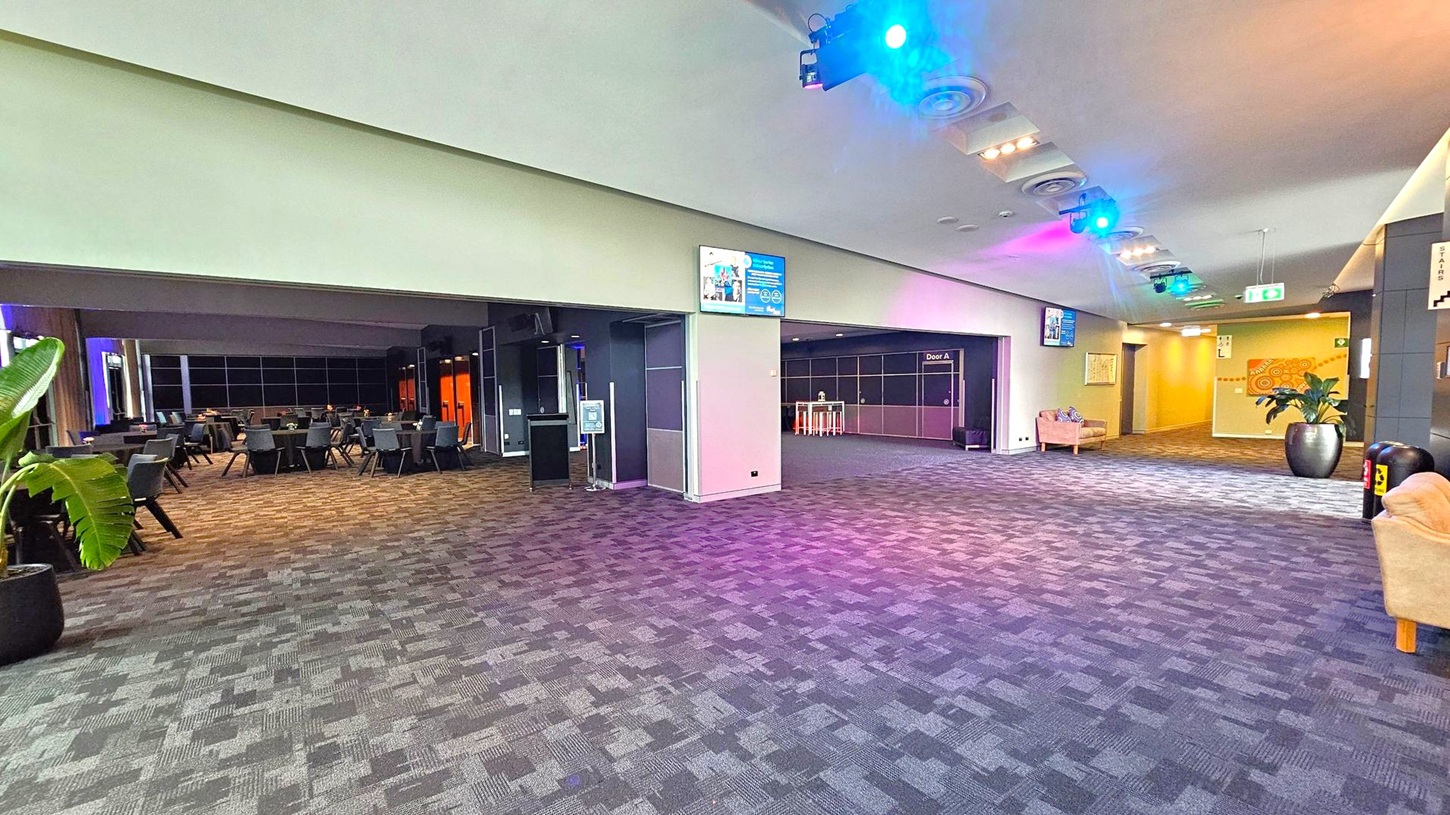 A spacious indoor area with a patterned carpet floor. On the left, there are tables and chairs arranged in a dining setup, with a large potted plant nearby. The right side features a hallway with a large potted plant and a beige sofa. The ceiling has recessed lighting and coloured spotlights. Digital screens and signs are mounted on the walls, and there are doors labelled "Door A". The walls are decorated with abstract art. A spacious indoor area with a patterned carpet floor. On the left, there are tables and chairs arranged in a dining setup, with a large potted plant nearby. The right side features a hallway with a large potted plant and a beige sofa. The ceiling has recessed lighting and coloured spotlights. Digital screens and signs are mounted on the walls, and there are doors labelled "Door A". The walls are decorated with abstract art.