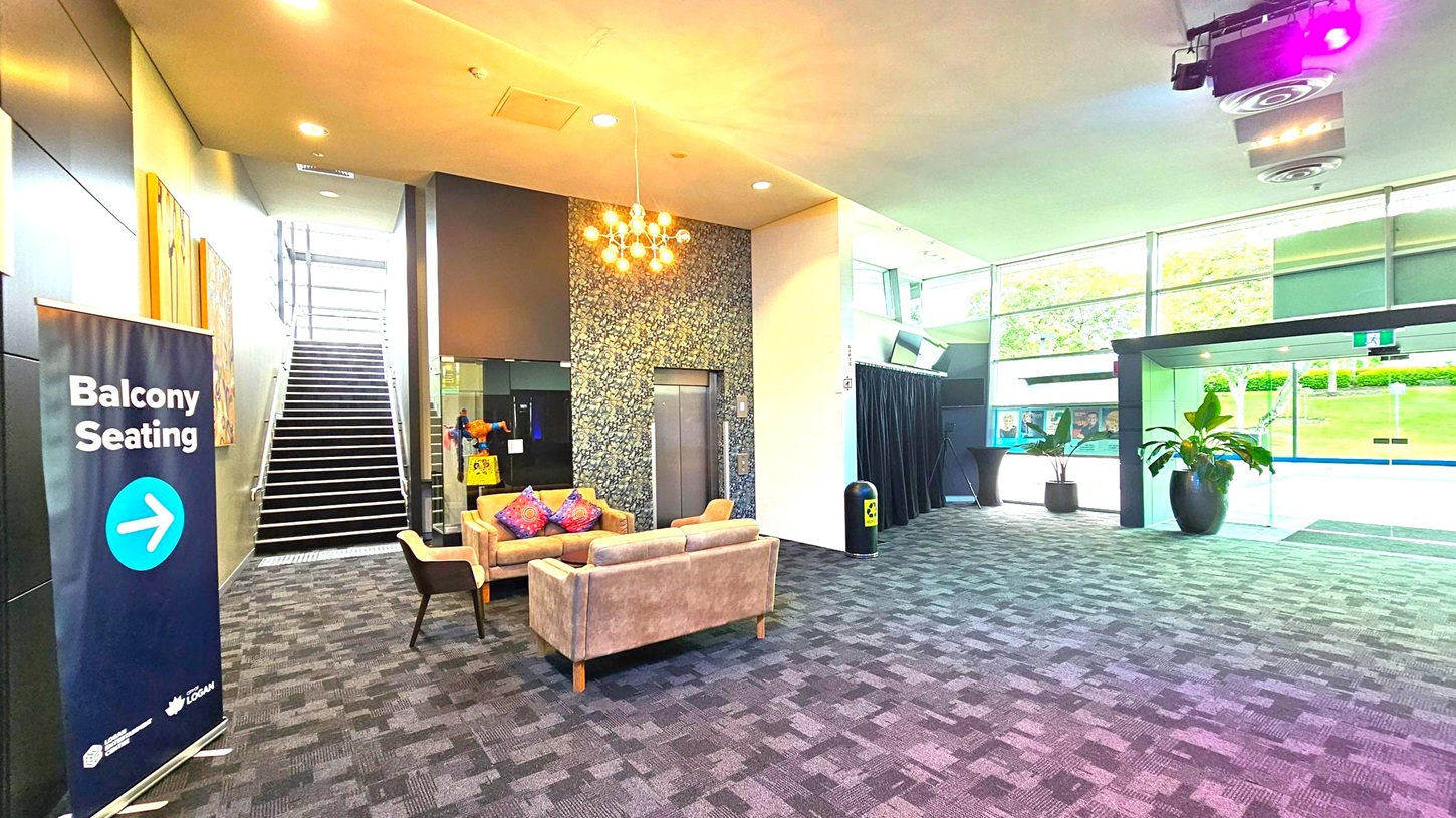 A modern interior space with a patterned carpet floor. On the left, a sign indicates "Balcony Seating" with an arrow. A staircase with metal railings leads upwards. In the centre, a seating area features a beige sofa and chairs with colourful cushions, under a cluster of hanging lights. An elevator is nearby. On the right, large windows provide a view of greenery outside. A potted plant and a small table are near the glass entrance doors. The ceiling has recessed lighting and a purple spotlight. A modern interior space with a patterned carpet floor. On the left, a sign indicates "Balcony Seating" with an arrow. A staircase with metal railings leads upwards. In the centre, a seating area features a beige sofa and chairs with colourful cushions, under a cluster of hanging lights. An elevator is nearby. On the right, large windows provide a view of greenery outside. A potted plant and a small table are near the glass entrance doors. The ceiling has recessed lighting and a purple spotlight.