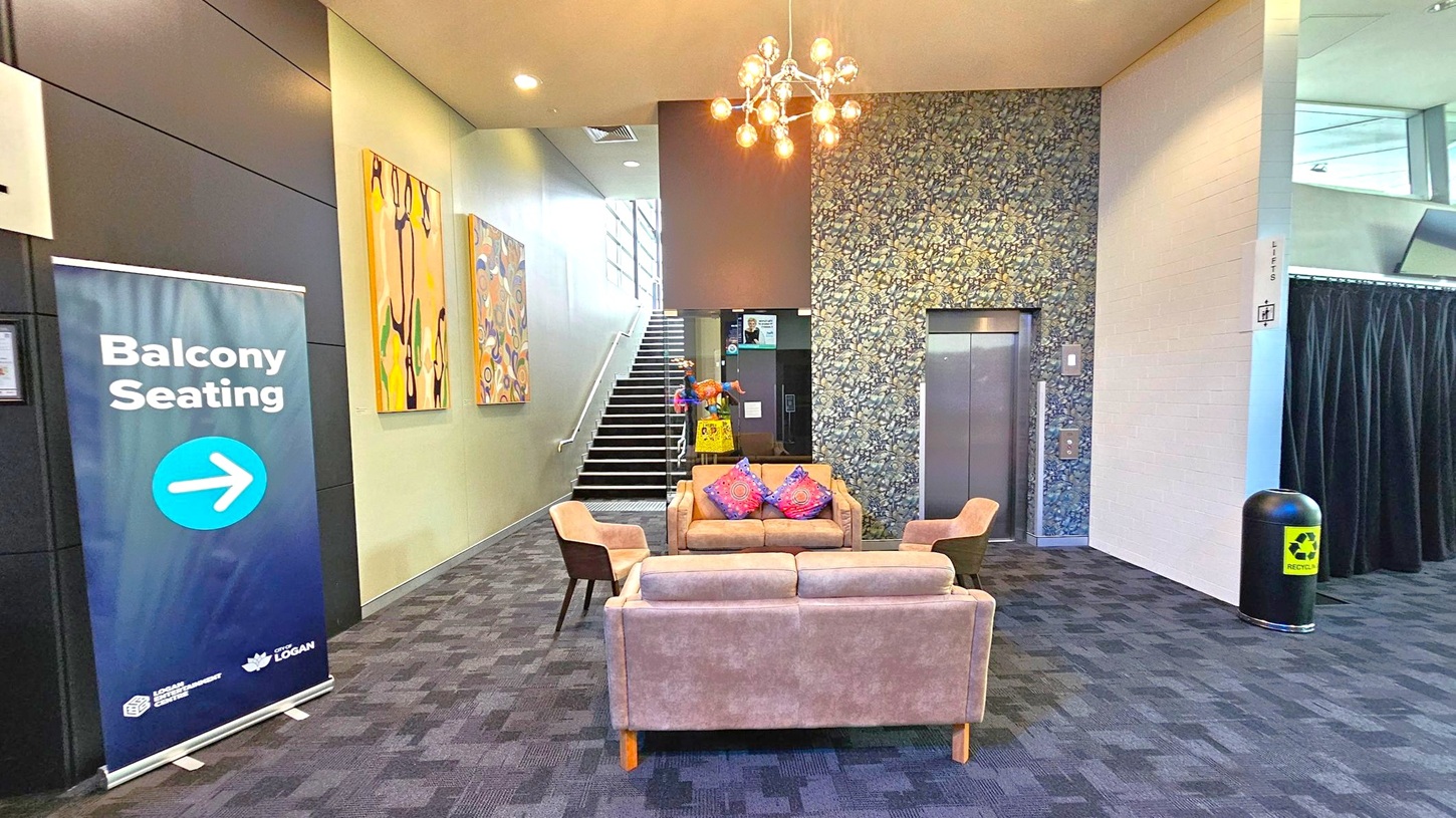 A modern interior space with a patterned carpeted floor. In the centre, there are two beige sofas and two chairs with colourful cushions. A chandelier hangs from the ceiling. On the left, a sign reads "Balcony Seating" with an arrow. Behind the seating area, a staircase leads upwards, flanked by abstract art on the wall. To the right, there's a lift with a floral-patterned wall and a recycling bin nearby. A modern interior space with a patterned carpeted floor. In the centre, there are two beige sofas and two chairs with colourful cushions. A chandelier hangs from the ceiling. On the left, a sign reads "Balcony Seating" with an arrow. Behind the seating area, a staircase leads upwards, flanked by abstract art on the wall. To the right, there's a lift with a floral-patterned wall and a recycling bin nearby.