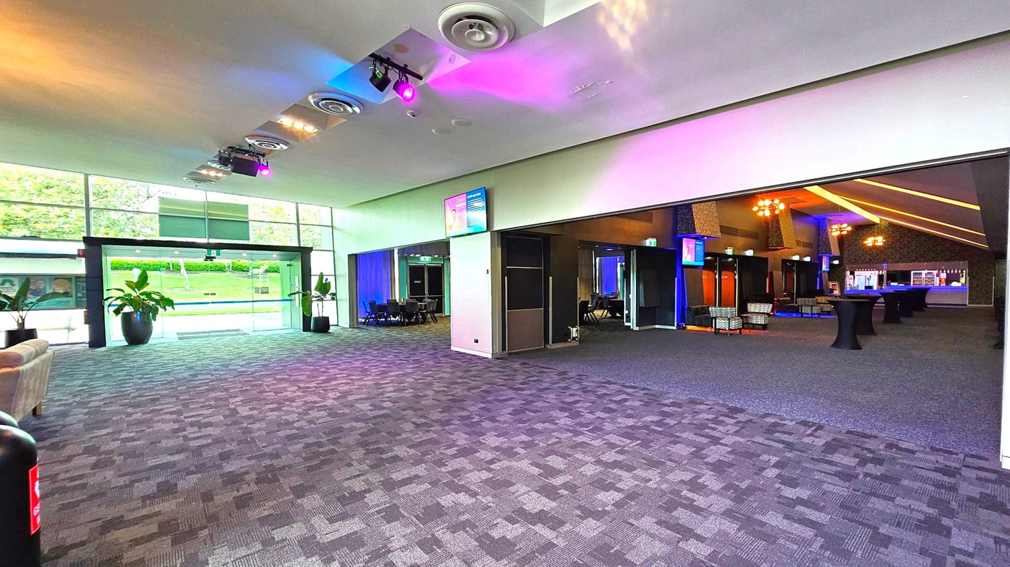 Spacious interior with a patterned carpet floor. On the left, large glass doors lead outside, flanked by potted plants. The ceiling features circular vents and colourful lights. On the right, an open area with tables and chairs, illuminated by warm lighting. A bar is visible in the background. Monitors are mounted on the walls, and a sofa is partially visible on the far left. Spacious interior with a patterned carpet floor. On the left, large glass doors lead outside, flanked by potted plants. The ceiling features circular vents and colourful lights. On the right, an open area with tables and chairs, illuminated by warm lighting. A bar is visible in the background. Monitors are mounted on the walls, and a sofa is partially visible on the far left.