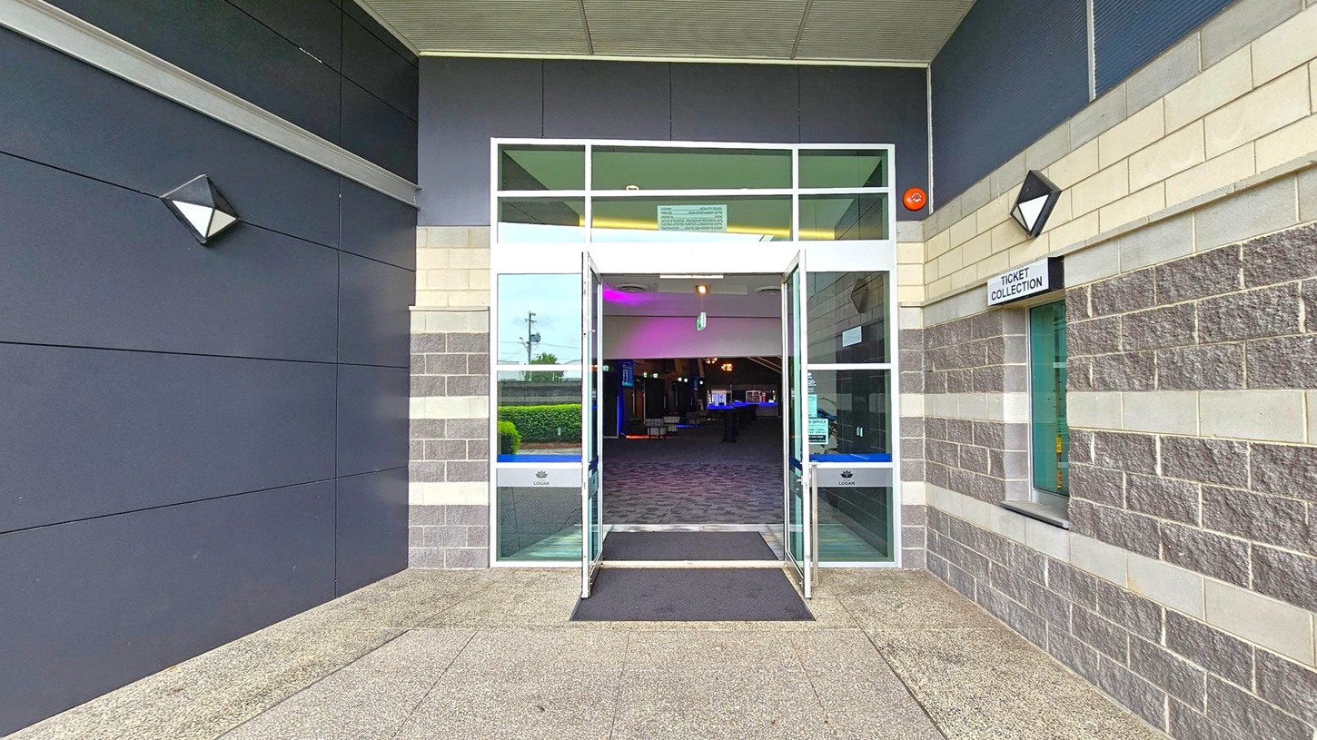 Entrance to a building with double glass doors open, leading to a dimly lit interior. The exterior walls are a mix of dark panels on the left and grey brick on the right. A sign above a window on the right reads "Ticket Collection". The floor is a speckled concrete, and there is a black mat in front of the doors. The ceiling is light-coloured with a grid pattern. Triangular wall lights are mounted on either side of the entrance. Entrance to a building with double glass doors open, leading to a dimly lit interior. The exterior walls are a mix of dark panels on the left and grey brick on the right. A sign above a window on the right reads "Ticket Collection". The floor is a speckled concrete, and there is a black mat in front of the doors. The ceiling is light-coloured with a grid pattern. Triangular wall lights are mounted on either side of the entrance.