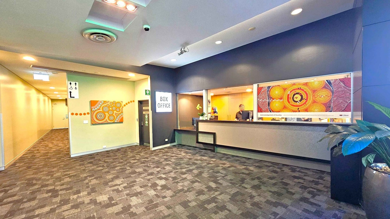 A modern interior with a box office on the right, featuring a counter and a person behind it. Above the counter is a colourful artwork. On the left, a hallway extends with a patterned carpet and a wall adorned with another piece of art. The ceiling has recessed lighting and a circular vent. A potted plant is in the foreground on the right. The floor is covered with a patterned carpet. A modern interior with a box office on the right, featuring a counter and a person behind it. Above the counter is a colourful artwork. On the left, a hallway extends with a patterned carpet and a wall adorned with another piece of art. The ceiling has recessed lighting and a circular vent. A potted plant is in the foreground on the right. The floor is covered with a patterned carpet.