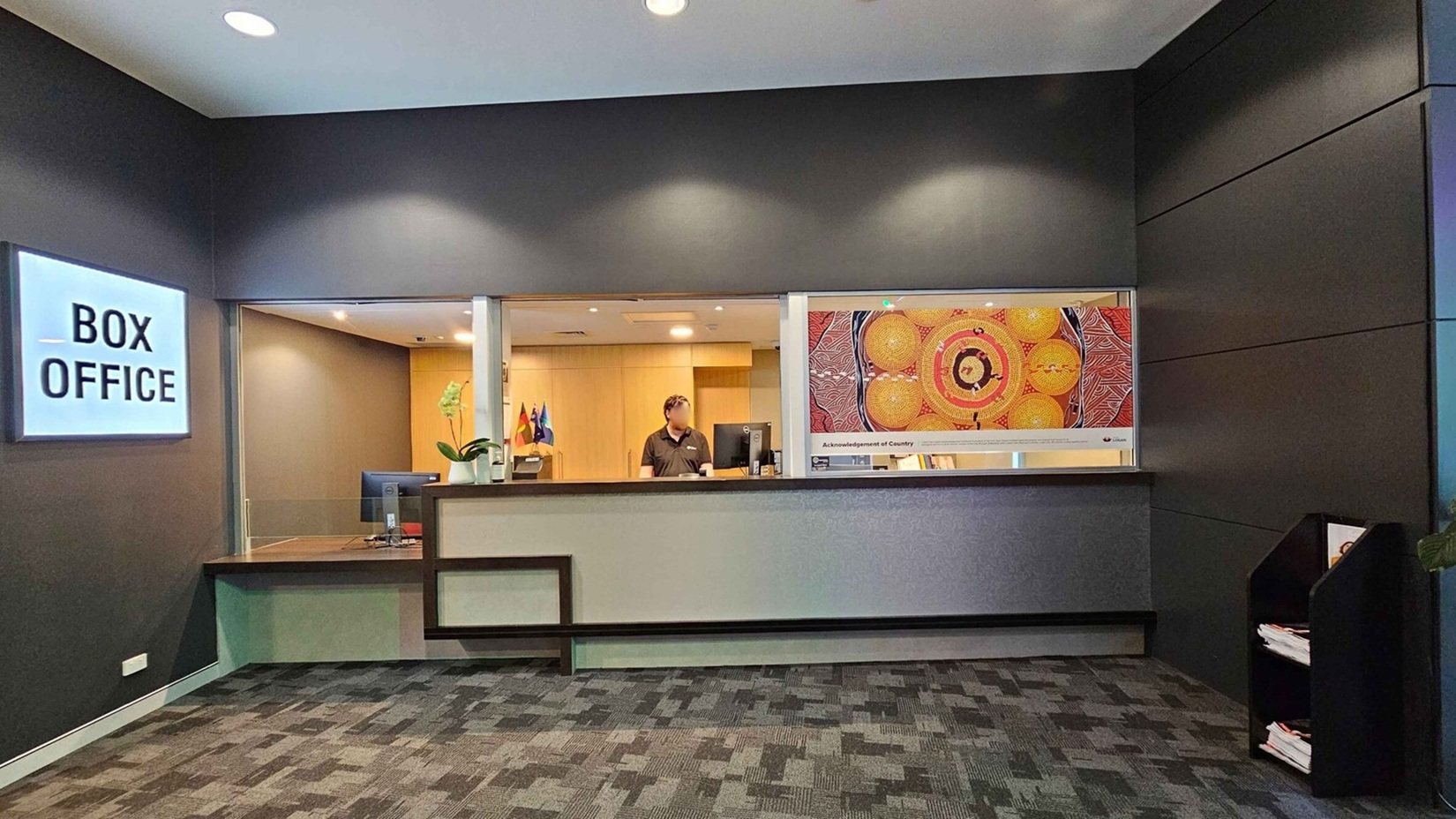 A box office area with a grey and black colour scheme. On the left, a lit sign reads "BOX OFFICE" above a counter with a computer and a potted plant. In the centre, a person stands behind the counter, with flags visible in the background. On the right, a colourful artwork is displayed on the wall. Below the artwork, a shelf holds brochures or papers. The floor is carpeted with a patterned design, and the ceiling has recessed lighting. A box office area with a grey and black colour scheme. On the left, a lit sign reads "BOX OFFICE" above a counter with a computer and a potted plant. In the centre, a person stands behind the counter, with flags visible in the background. On the right, a colourful artwork is displayed on the wall. Below the artwork, a shelf holds brochures or papers. The floor is carpeted with a patterned design, and the ceiling has recessed lighting.