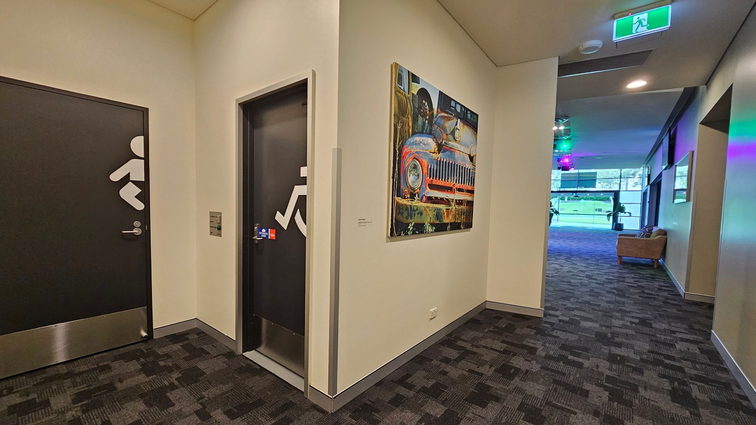A hallway with two black doors on the left, each marked with white symbols indicating accessibility. Between the doors, a colourful painting of an old car is mounted on the wall. The floor is covered with a patterned carpet. To the right, the hallway opens into a larger room with a sofa and large windows. The ceiling has recessed lighting and an illuminated green exit sign. A hallway with two black doors on the left, each marked with white symbols indicating accessibility. Between the doors, a colourful painting of an old car is mounted on the wall. The floor is covered with a patterned carpet. To the right, the hallway opens into a larger room with a sofa and large windows. The ceiling has recessed lighting and an illuminated green exit sign.