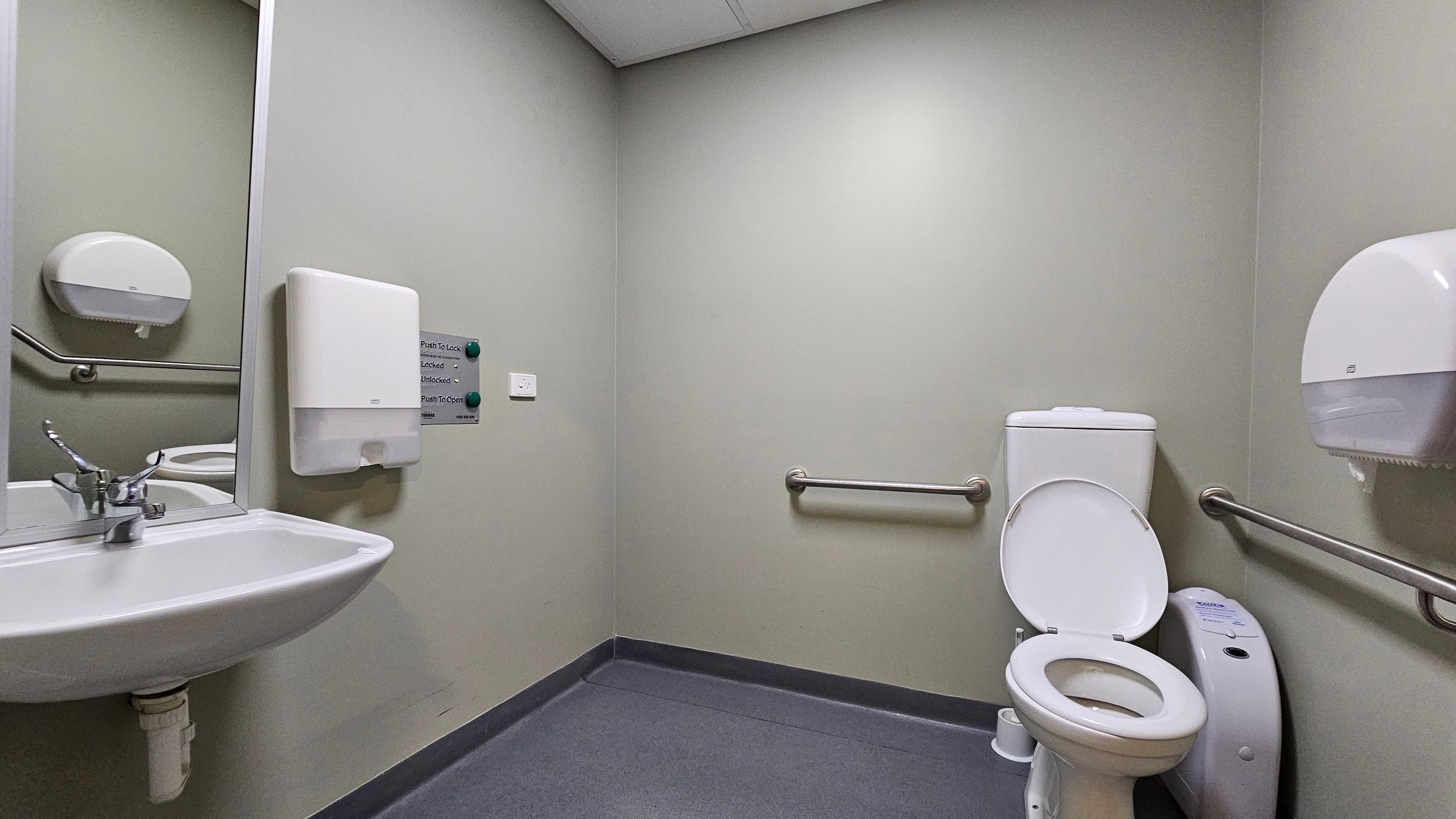 A public restroom with grey walls and a light grey ceiling. On the left, a white sink with a metal tap is mounted below a large mirror. A paper towel dispenser is above the sink. In the centre, a wall-mounted panel with buttons for locking and unlocking. On the right, a white toilet with the lid open, flanked by metal grab bars. A toilet paper dispenser is mounted on the wall. The floor is covered with dark grey tiles. A public restroom with grey walls and a light grey ceiling. On the left, a white sink with a metal tap is mounted below a large mirror. A paper towel dispenser is above the sink. In the centre, a wall-mounted panel with buttons for locking and unlocking. On the right, a white toilet with the lid open, flanked by metal grab bars. A toilet paper dispenser is mounted on the wall. The floor is covered with dark grey tiles.