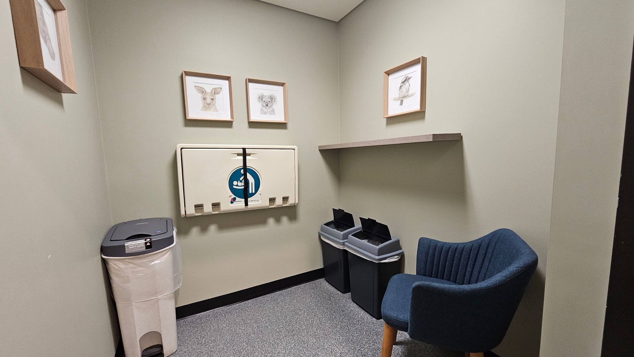 A small room with light green walls. On the left, a beige nappy changing station is mounted on the wall, with a waste bin below it. Above the station are three framed animal sketches. On the right, a wooden shelf is mounted on the wall, with two black bins beneath it. A blue upholstered chair with wooden legs is positioned in the corner. The floor is covered with grey speckled carpet tiles. A small room with light green walls. On the left, a beige nappy changing station is mounted on the wall, with a waste bin below it. Above the station are three framed animal sketches. On the right, a wooden shelf is mounted on the wall, with two black bins beneath it. A blue upholstered chair with wooden legs is positioned in the corner. The floor is covered with grey speckled carpet tiles.