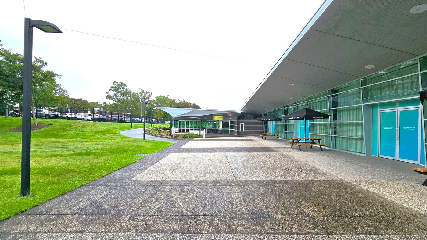 A modern building with a glass facade on the right, featuring blue doors and large windows. Two picnic tables with black umbrellas are placed along the building. The foreground has a paved area leading to a grassy lawn on the left, with a path and several trees. A row of parked cars is visible in the background, separated by a fence. The ceiling of the building extends outward, providing some shelter over the entrance area. A modern building with a glass facade on the right, featuring blue doors and large windows. Two picnic tables with black umbrellas are placed along the building. The foreground has a paved area leading to a grassy lawn on the left, with a path and several trees. A row of parked cars is visible in the background, separated by a fence. The ceiling of the building extends outward, providing some shelter over the entrance area.