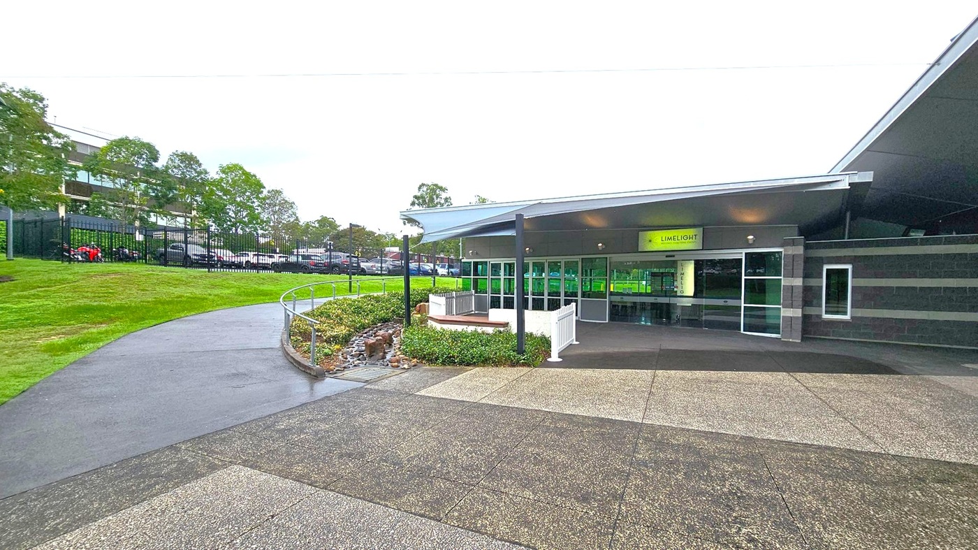 A modern building with a sign reading "LIMELIGHT" above the entrance, featuring large glass doors and windows. The structure has a sloped roof and is surrounded by a paved area with a mix of grey and green tiles. To the left, a curved path leads up a grassy hill, bordered by a metal railing and small shrubs. In the background, there are trees and a fenced parking area with several cars. A modern building with a sign reading "LIMELIGHT" above the entrance, featuring large glass doors and windows. The structure has a sloped roof and is surrounded by a paved area with a mix of grey and green tiles. To the left, a curved path leads up a grassy hill, bordered by a metal railing and small shrubs. In the background, there are trees and a fenced parking area with several cars.