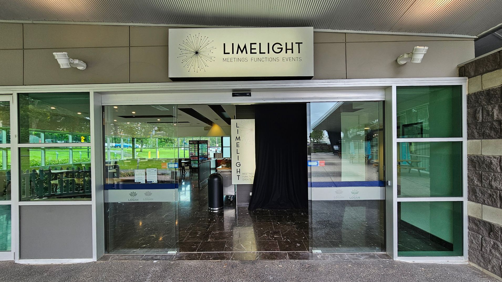 A glass entrance with a sign above reading "LIMELIGHT MEETINGS FUNCTIONS EVENTS". The entrance has large glass doors and windows with green-tinted panels on either side. Inside, there is a black curtain and a glimpse of a seating area with tables and chairs. The floor is dark and glossy, reflecting the interior lights. The exterior has a textured stone wall on the right and a grey ceiling with lights. A glass entrance with a sign above reading "LIMELIGHT MEETINGS FUNCTIONS EVENTS". The entrance has large glass doors and windows with green-tinted panels on either side. Inside, there is a black curtain and a glimpse of a seating area with tables and chairs. The floor is dark and glossy, reflecting the interior lights. The exterior has a textured stone wall on the right and a grey ceiling with lights.