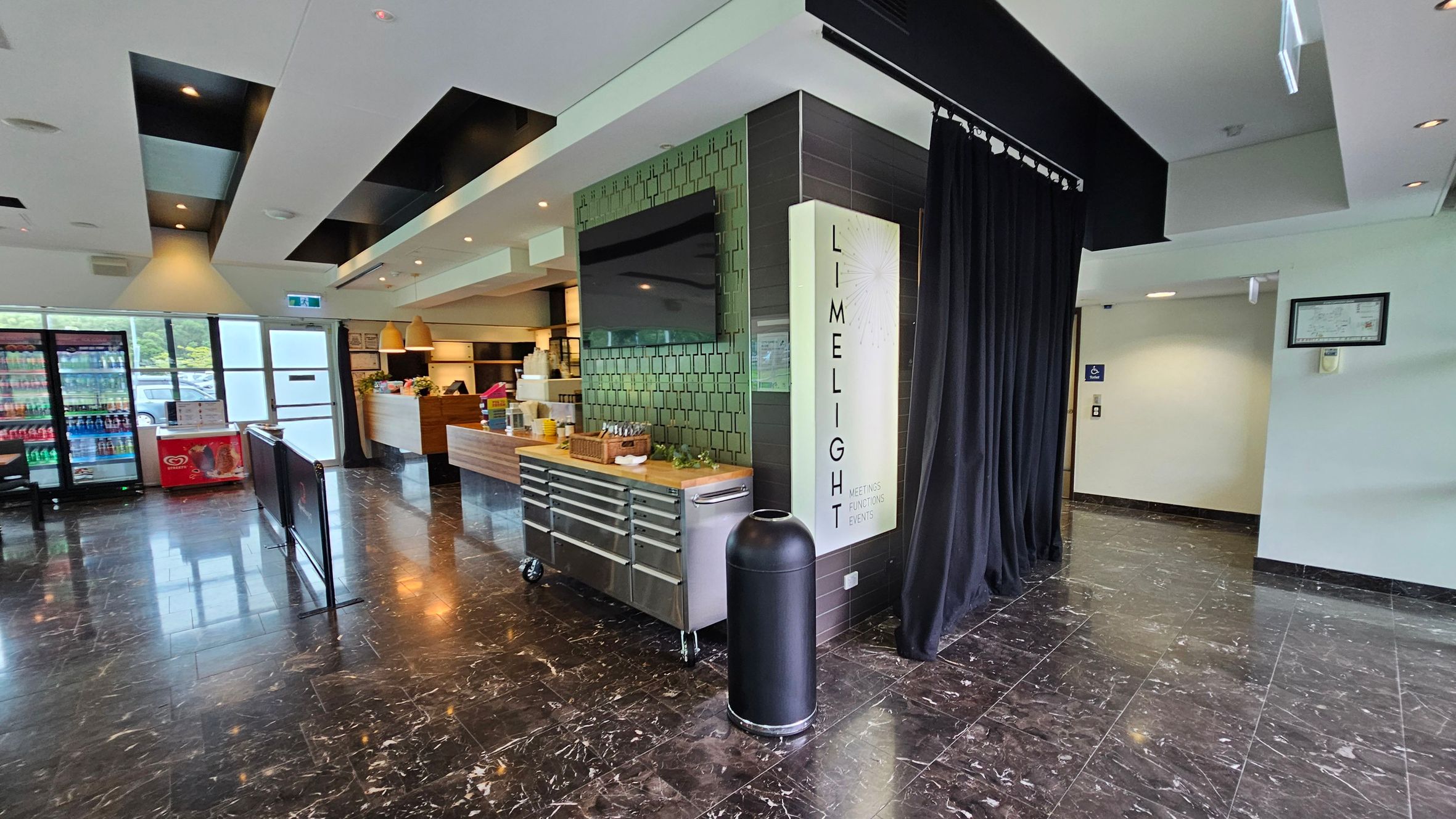 A modern interior with a glossy black marble floor. On the left, there are large windows and a fridge stocked with drinks. In the centre, a counter with wooden accents and hanging lights is visible. A black curtain and a sign reading "LIMELIGHT" are on the right, next to a black cylindrical bin. The ceiling features recessed lighting and black panels. A modern interior with a glossy black marble floor. On the left, there are large windows and a fridge stocked with drinks. In the centre, a counter with wooden accents and hanging lights is visible. A black curtain and a sign reading "LIMELIGHT" are on the right, next to a black cylindrical bin. The ceiling features recessed lighting and black panels.