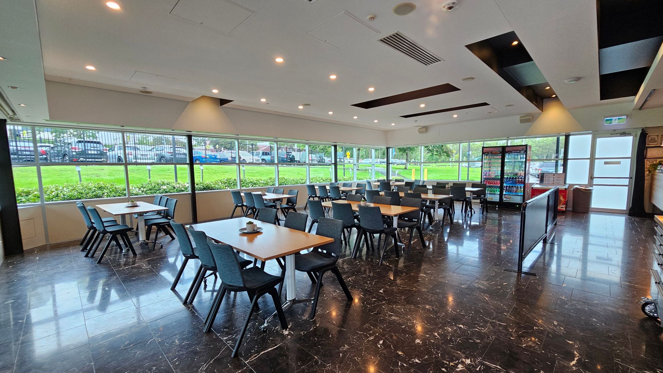 A spacious dining area with several wooden tables and black chairs arranged neatly. Large windows on the left and background allow natural light to illuminate the room, offering a view of a grassy area and parked cars outside. On the right, there are refrigerated display units stocked with beverages. The floor is dark with a polished finish, and the ceiling features recessed lighting and air vents. A spacious dining area with several wooden tables and black chairs arranged neatly. Large windows on the left and background allow natural light to illuminate the room, offering a view of a grassy area and parked cars outside. On the right, there are refrigerated display units stocked with beverages. The floor is dark with a polished finish, and the ceiling features recessed lighting and air vents.