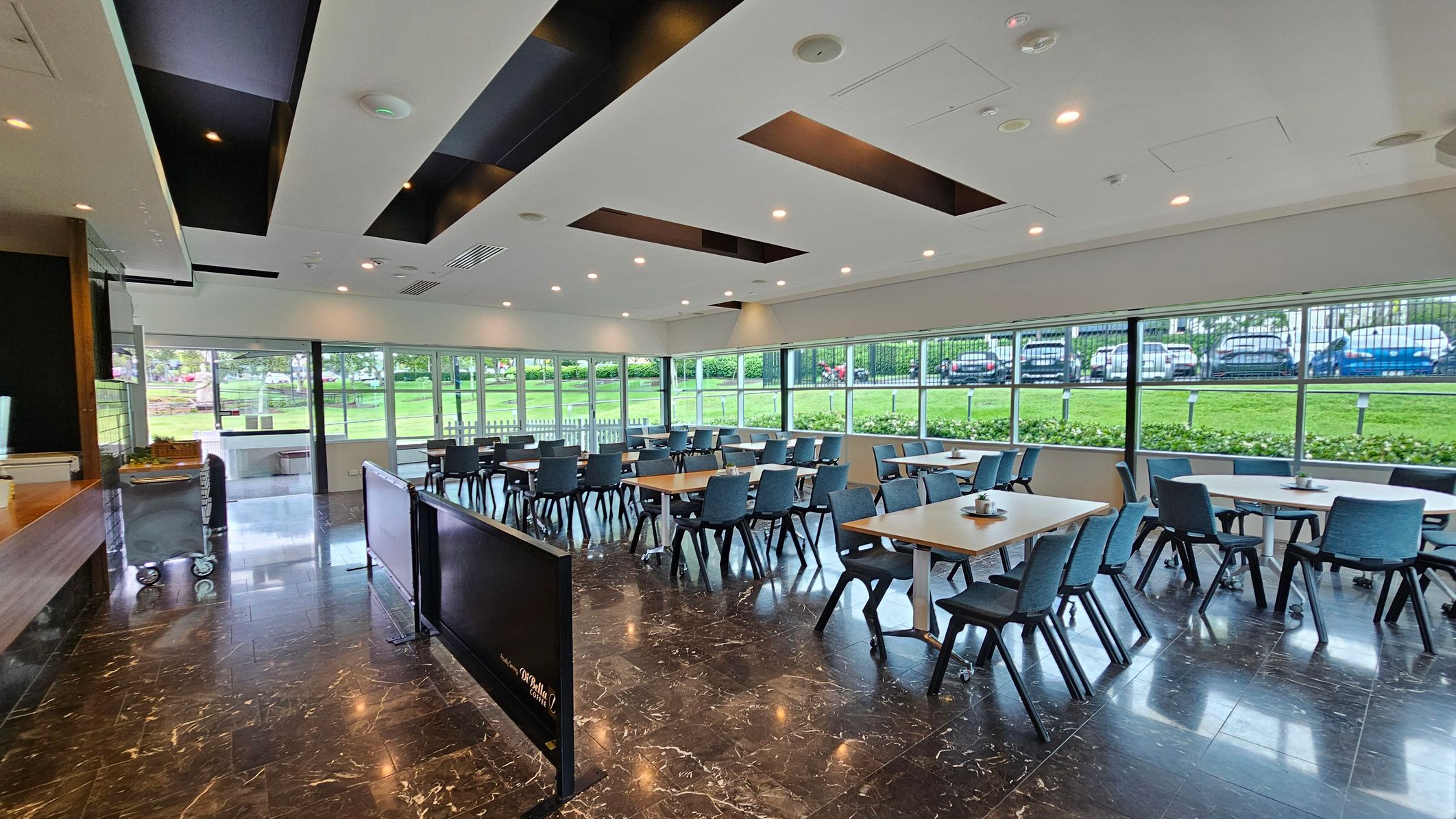 A spacious dining area with dark marble flooring and a modern ceiling featuring recessed lighting and black accents. On the left, there's a counter with a trolley nearby. The right side has several tables and chairs arranged neatly. Large windows in the background offer a view of a grassy area and parked cars outside. A spacious dining area with dark marble flooring and a modern ceiling featuring recessed lighting and black accents. On the left, there's a counter with a trolley nearby. The right side has several tables and chairs arranged neatly. Large windows in the background offer a view of a grassy area and parked cars outside.
