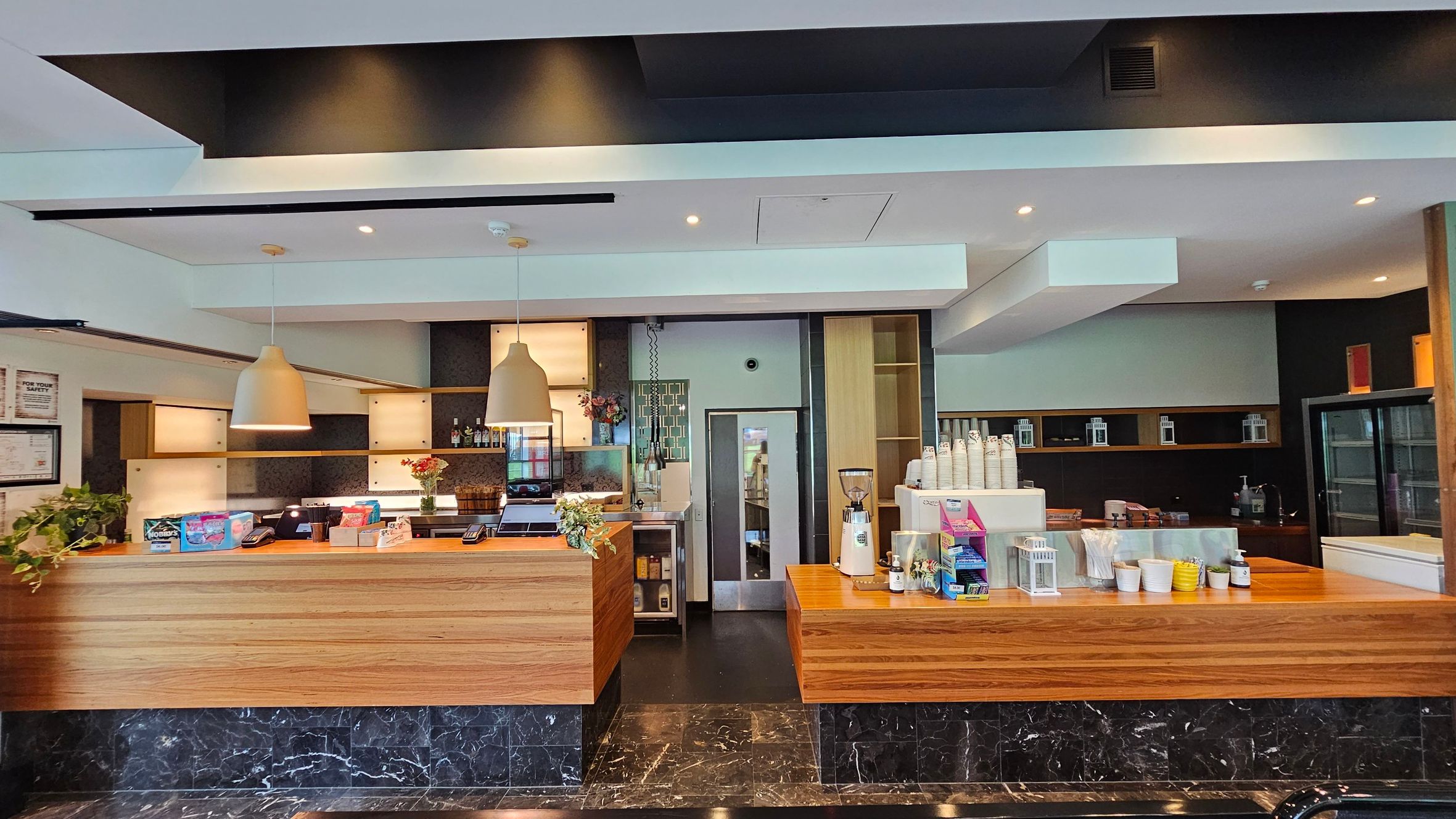 A modern café counter with a wooden finish. On the left, there are two large pendant lights above the counter, which displays snacks and a cash register. A plant and some flowers add a touch of greenery. In the centre, a door leads to the back area. On the right, a coffee machine and cups are visible, with shelves holding various items. The floor is dark marble, and the ceiling is white with recessed lighting. A modern café counter with a wooden finish. On the left, there are two large pendant lights above the counter, which displays snacks and a cash register. A plant and some flowers add a touch of greenery. In the centre, a door leads to the back area. On the right, a coffee machine and cups are visible, with shelves holding various items. The floor is dark marble, and the ceiling is white with recessed lighting.