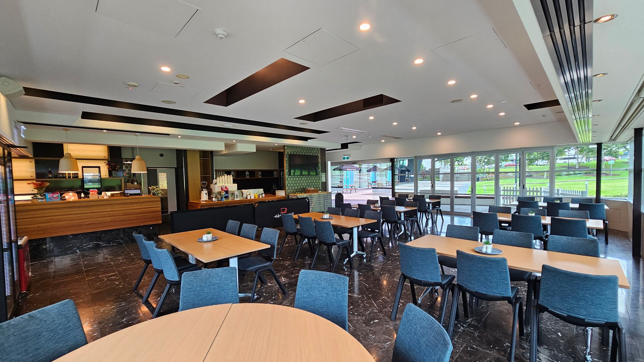 A spacious dining area with several wooden tables and blue chairs arranged neatly. On the left, there's a counter with pendant lights above, displaying various items and a coffee machine. The floor is dark marble, and the ceiling is white with recessed lighting. Large windows on the right offer a view of a green outdoor area with trees and picnic tables. The room is well-lit and modern in design. A spacious dining area with several wooden tables and blue chairs arranged neatly. On the left, there's a counter with pendant lights above, displaying various items and a coffee machine. The floor is dark marble, and the ceiling is white with recessed lighting. Large windows on the right offer a view of a green outdoor area with trees and picnic tables. The room is well-lit and modern in design.