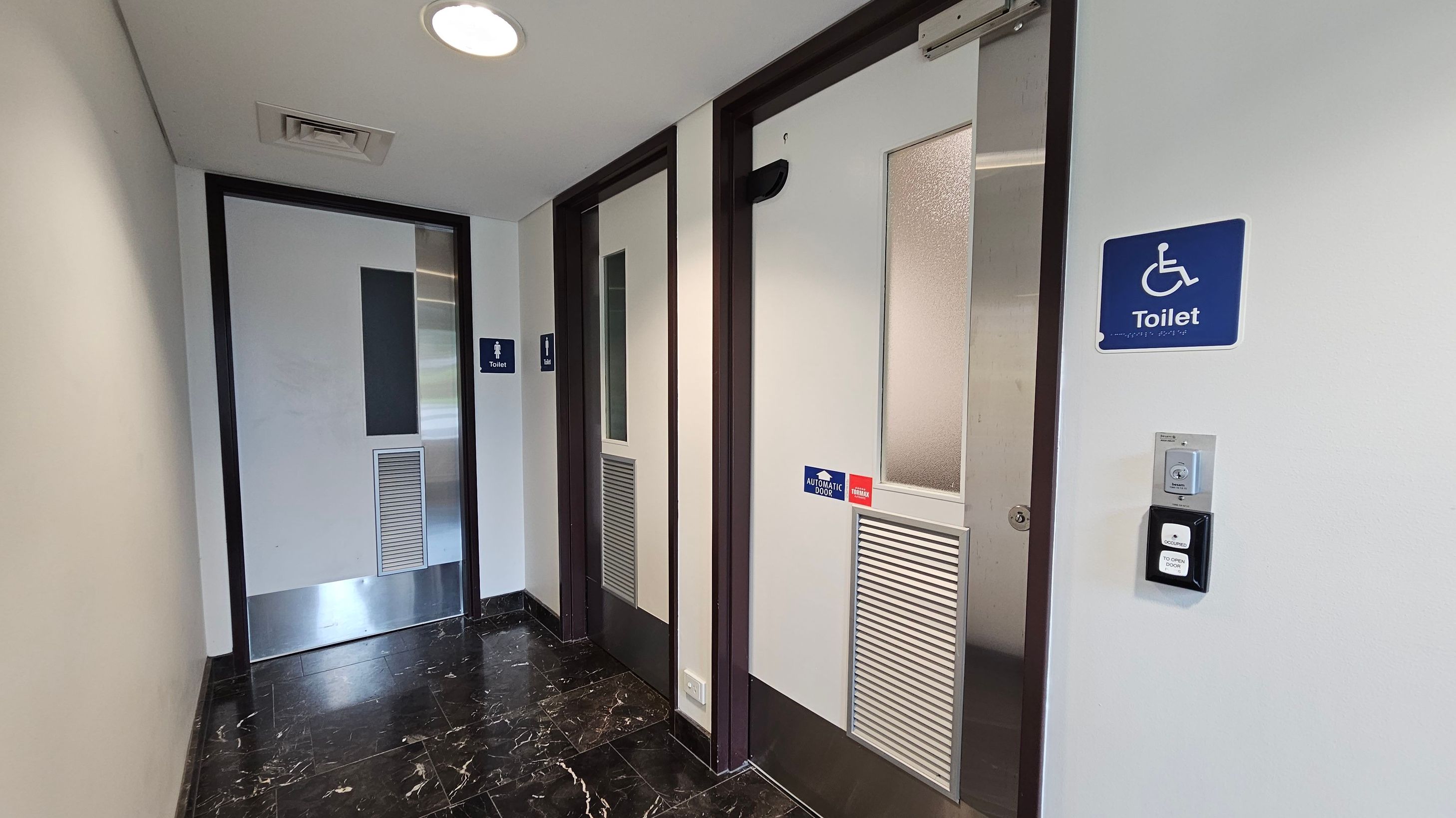 A corridor with two toilet doors on the right, one marked with a wheelchair symbol for accessibility. Both doors have frosted glass panels and ventilation grilles. The accessible toilet has an automatic door sign and a button for operation. The floor is black marble with white veining, and the ceiling is white with recessed lighting and a vent. On the left, a plain wall leads to another door at the end of the corridor. A corridor with two toilet doors on the right, one marked with a wheelchair symbol for accessibility. Both doors have frosted glass panels and ventilation grilles. The accessible toilet has an automatic door sign and a button for operation. The floor is black marble with white veining, and the ceiling is white with recessed lighting and a vent. On the left, a plain wall leads to another door at the end of the corridor.