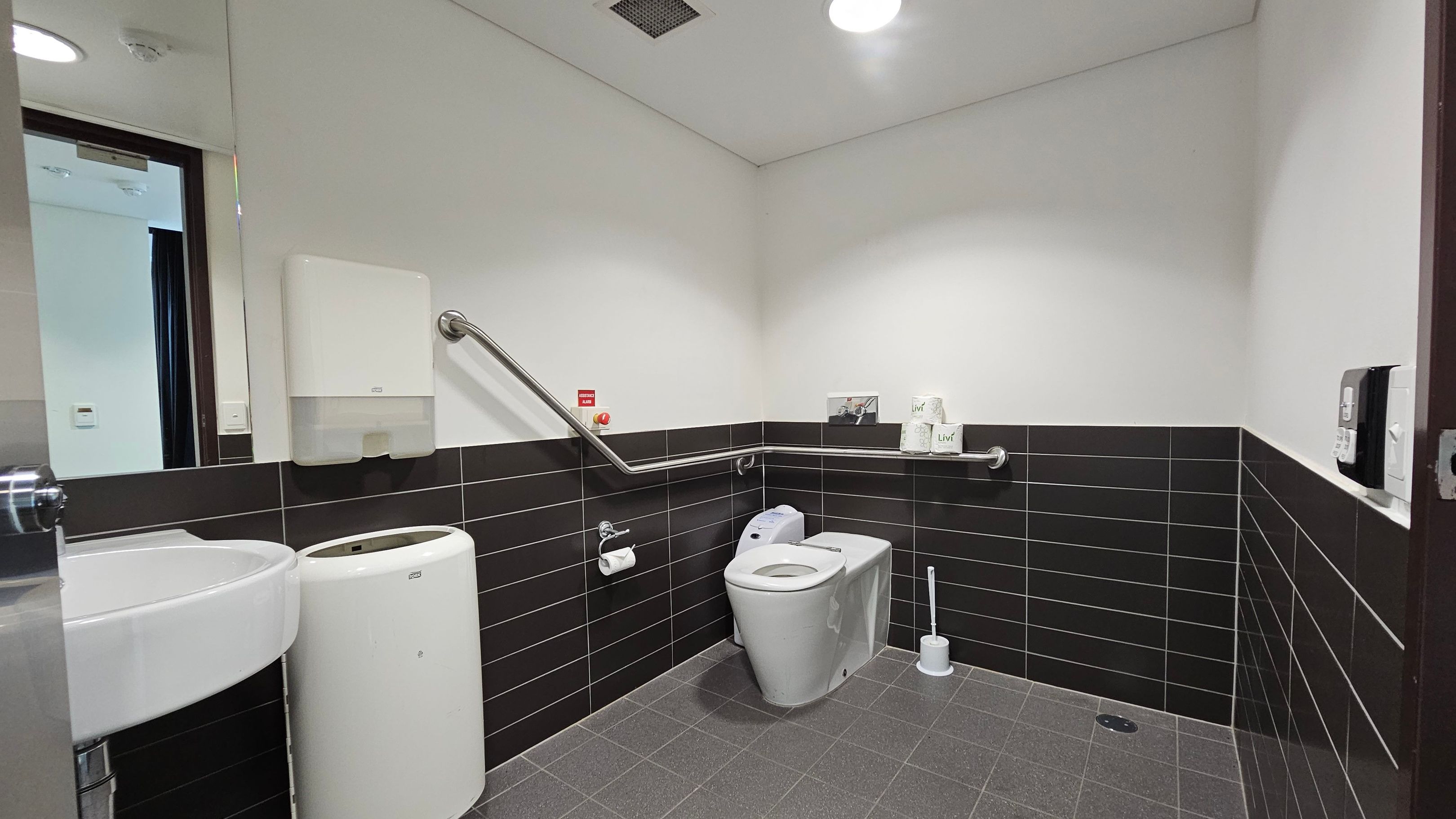 Accessible bathroom with grey tiled floor and white ceiling. On the left, a white sink with a mirror above and a bin beside it. The middle features a toilet with a support rail, toilet paper, and a brush. The walls are half-tiled with black tiles. A paper towel dispenser and emergency button are mounted on the wall. On the right, a soap dispenser is visible. The room is well-lit with a ceiling light. Accessible bathroom with grey tiled floor and white ceiling. On the left, a white sink with a mirror above and a bin beside it. The middle features a toilet with a support rail, toilet paper, and a brush. The walls are half-tiled with black tiles. A paper towel dispenser and emergency button are mounted on the wall. On the right, a soap dispenser is visible. The room is well-lit with a ceiling light.