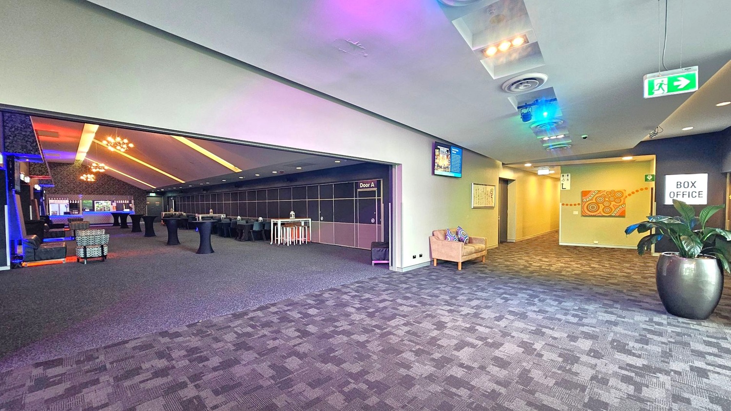 A spacious interior with a patterned carpeted floor. On the left, an open area with high tables and chairs, illuminated by modern chandeliers. A sign reads "Door A". On the right, a hallway with a box office sign, a potted plant, and a beige sofa with colourful cushions. The ceiling features recessed lighting and air vents. An exit sign is visible above the hallway. A spacious interior with a patterned carpeted floor. On the left, an open area with high tables and chairs, illuminated by modern chandeliers. A sign reads "Door A". On the right, a hallway with a box office sign, a potted plant, and a beige sofa with colourful cushions. The ceiling features recessed lighting and air vents. An exit sign is visible above the hallway.