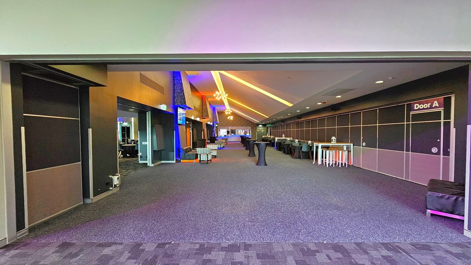 A spacious interior with a high, slanted ceiling featuring recessed lighting and chandeliers. The floor is carpeted in a dark grey pattern. On the left, there are open doorways and seating areas with armchairs. On the right, a long wall with a sign for "Door A" and several high tables with stools. In the background, a bar area is visible, and the space is illuminated with colourful lighting accents. A spacious interior with a high, slanted ceiling featuring recessed lighting and chandeliers. The floor is carpeted in a dark grey pattern. On the left, there are open doorways and seating areas with armchairs. On the right, a long wall with a sign for "Door A" and several high tables with stools. In the background, a bar area is visible, and the space is illuminated with colourful lighting accents.