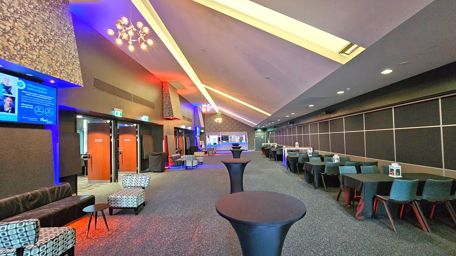 A spacious, modern lounge area with a high, slanted ceiling featuring recessed lighting. On the left, there are patterned chairs and a black leather sofa near wooden doors. A digital screen is mounted on the wall. The right side has a row of tables with teal chairs, each adorned with a small decorative item. The floor is carpeted, and several tall, round cocktail tables are positioned along the centre. The lighting creates a warm, inviting atmosphere with blue and red accents. A spacious, modern lounge area with a high, slanted ceiling featuring recessed lighting. On the left, there are patterned chairs and a black leather sofa near wooden doors. A digital screen is mounted on the wall. The right side has a row of tables with teal chairs, each adorned with a small decorative item. The floor is carpeted, and several tall, round cocktail tables are positioned along the centre. The lighting creates a warm, inviting atmosphere with blue and red accents.