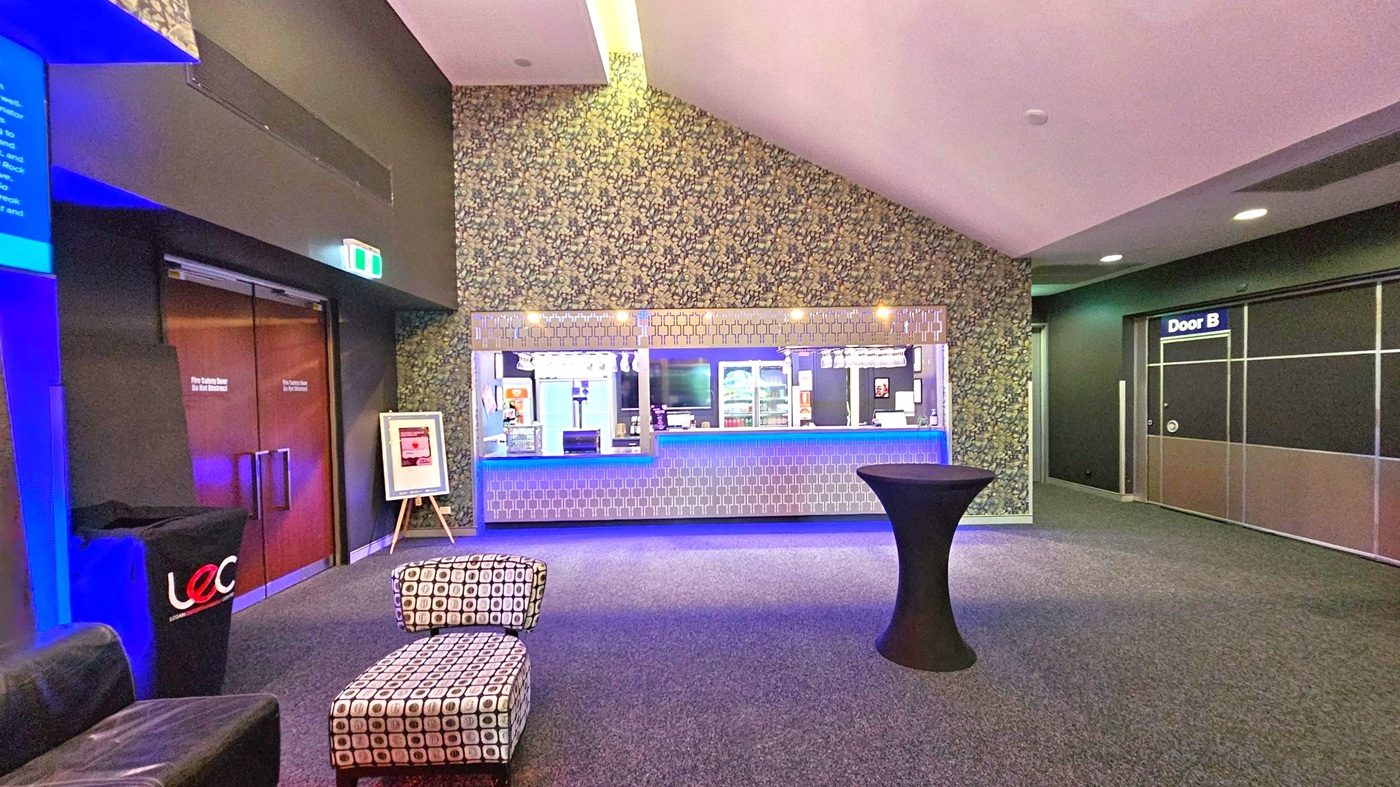 A modern interior with a patterned wall in the centre featuring a counter with a geometric design and blue lighting. Above the counter, glasses hang upside down. To the left, there are double wooden doors with a framed sign on an easel nearby. A patterned ottoman and a black cocktail table are in the foreground. On the right, a door labelled "Door B" is visible. The floor is carpeted, and the ceiling has recessed lighting. A modern interior with a patterned wall in the centre featuring a counter with a geometric design and blue lighting. Above the counter, glasses hang upside down. To the left, there are double wooden doors with a framed sign on an easel nearby. A patterned ottoman and a black cocktail table are in the foreground. On the right, a door labelled "Door B" is visible. The floor is carpeted, and the ceiling has recessed lighting.