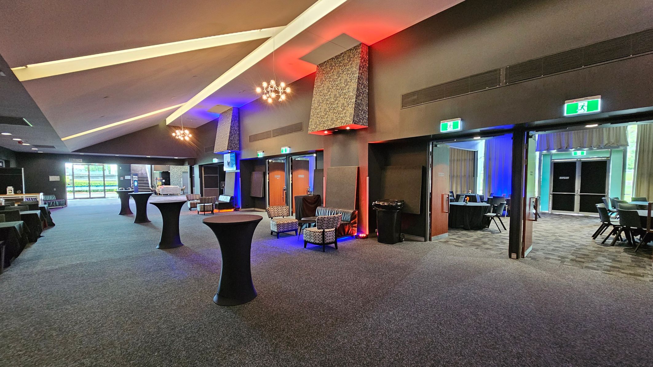 A spacious interior with a high, angled ceiling featuring modern light fixtures. The floor is carpeted in a grey tone. On the left, there are tables covered with black cloths and a large window with a view of greenery. In the centre, tall cocktail tables are scattered, and a seating area with patterned chairs is visible. On the right, there are open doors leading to another room with more tables and chairs. The walls are adorned with decorative panels and illuminated with coloured lights. A spacious interior with a high, angled ceiling featuring modern light fixtures. The floor is carpeted in a grey tone. On the left, there are tables covered with black cloths and a large window with a view of greenery. In the centre, tall cocktail tables are scattered, and a seating area with patterned chairs is visible. On the right, there are open doors leading to another room with more tables and chairs. The walls are adorned with decorative panels and illuminated with coloured lights.