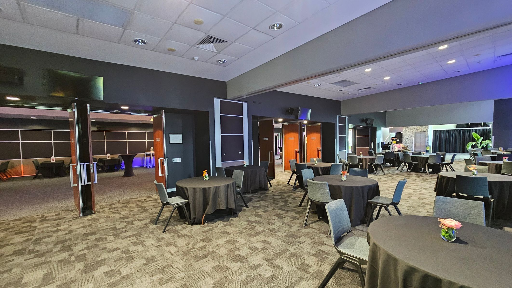 A spacious conference room with a patterned carpeted floor and a white panelled ceiling. Round tables covered with black tablecloths are scattered throughout, each adorned with a small floral centrepiece. Grey chairs are arranged around the tables. In the background, open double doors lead to another room with more tables and a blue-lit area. Large plants and a black curtain are visible in the far right corner. A spacious conference room with a patterned carpeted floor and a white panelled ceiling. Round tables covered with black tablecloths are scattered throughout, each adorned with a small floral centrepiece. Grey chairs are arranged around the tables. In the background, open double doors lead to another room with more tables and a blue-lit area. Large plants and a black curtain are visible in the far right corner.