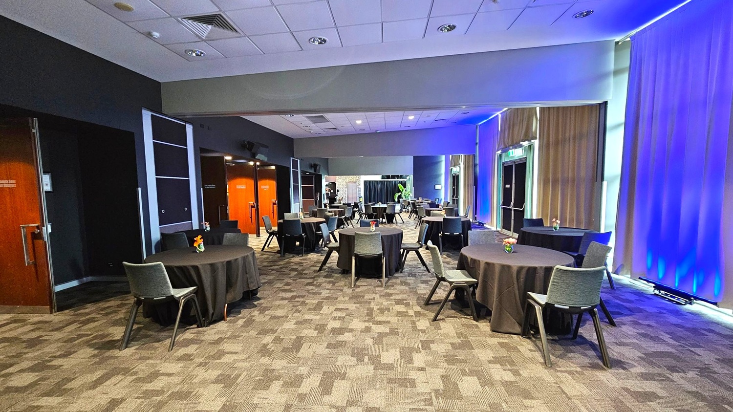 A conference room with round tables covered in black tablecloths, each with a small floral centrepiece. Grey chairs are arranged around the tables. The left side has wooden doors and dark walls, while the right side features large windows with beige curtains illuminated by blue lighting. The ceiling is white with recessed lighting, and the floor is carpeted in a patterned design. In the background, there is a plant and more seating. A conference room with round tables covered in black tablecloths, each with a small floral centrepiece. Grey chairs are arranged around the tables. The left side has wooden doors and dark walls, while the right side features large windows with beige curtains illuminated by blue lighting. The ceiling is white with recessed lighting, and the floor is carpeted in a patterned design. In the background, there is a plant and more seating.