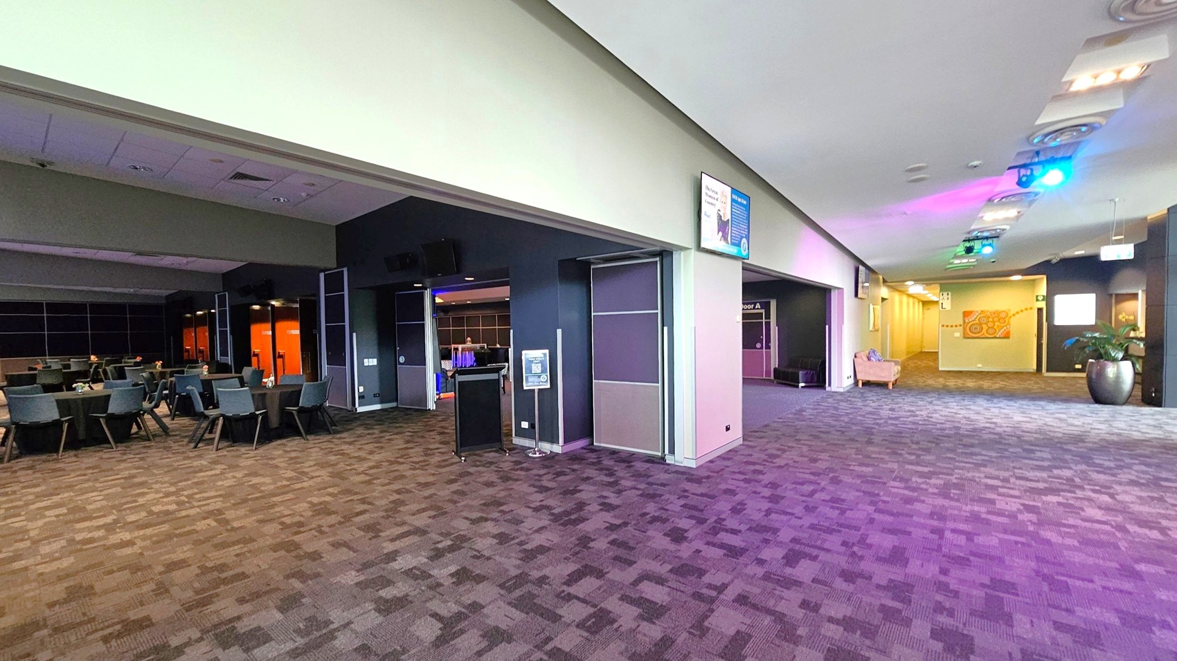 A spacious interior with a patterned carpet floor. On the left, there are tables and chairs arranged in a dining or conference style. The right side features a wide corridor with a potted plant and a beige armchair. The ceiling has recessed lighting and colourful spotlights. Walls are adorned with signs and a decorative mural. The area appears to be a multifunctional space, possibly in a conference centre or hotel. A spacious interior with a patterned carpet floor. On the left, there are tables and chairs arranged in a dining or conference style. The right side features a wide corridor with a potted plant and a beige armchair. The ceiling has recessed lighting and colourful spotlights. Walls are adorned with signs and a decorative mural. The area appears to be a multifunctional space, possibly in a conference centre or hotel.