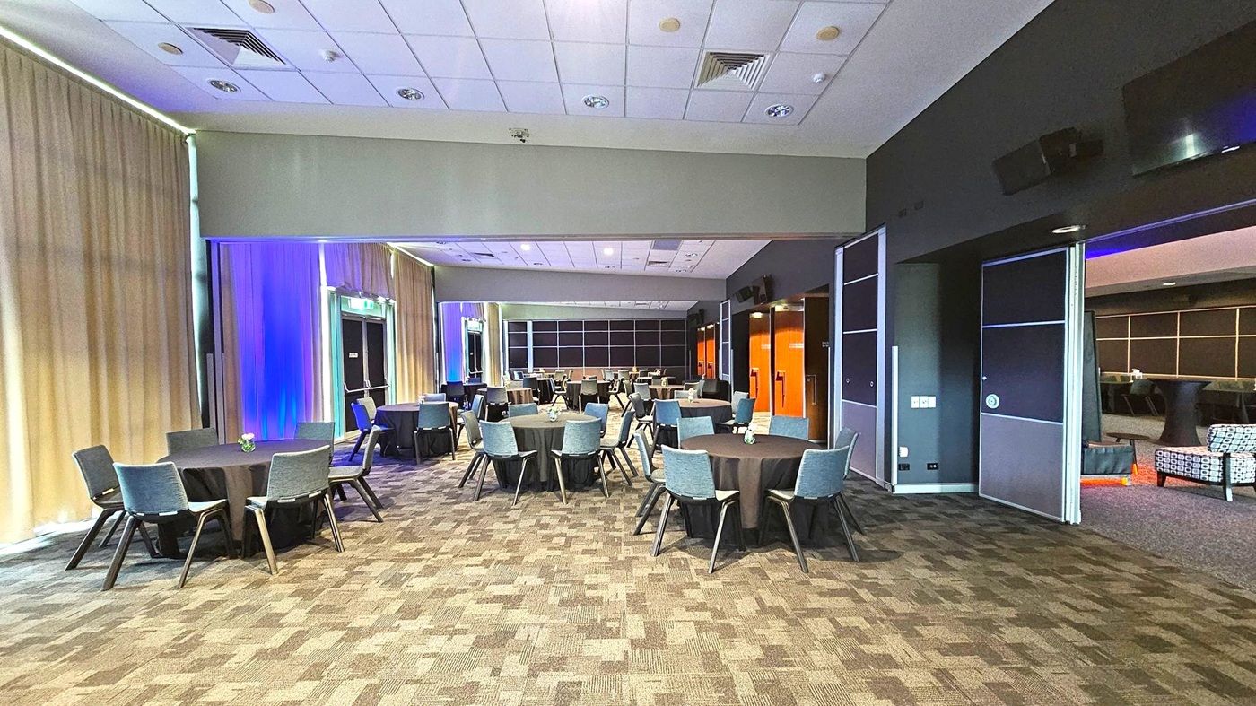 A spacious conference room with round tables covered in black tablecloths, each surrounded by grey chairs. The left side features beige curtains with blue lighting accents. The right side has dark walls with open sliding doors leading to another area with patterned chairs. The ceiling is white with recessed lighting and air vents. The floor is carpeted in a geometric pattern of brown and grey tones. A spacious conference room with round tables covered in black tablecloths, each surrounded by grey chairs. The left side features beige curtains with blue lighting accents. The right side has dark walls with open sliding doors leading to another area with patterned chairs. The ceiling is white with recessed lighting and air vents. The floor is carpeted in a geometric pattern of brown and grey tones.