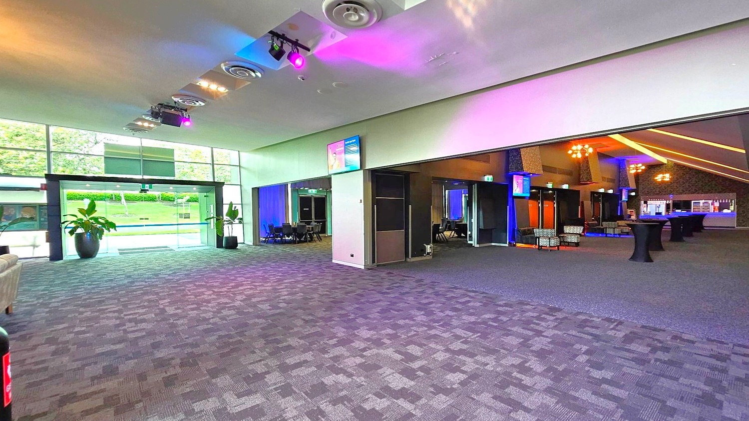 A spacious interior with a patterned carpeted floor. On the left, large glass doors lead outside, flanked by potted plants. The ceiling features circular vents and colourful lights. On the right, an open area with tables and chairs, illuminated by warm lighting. A bar is visible in the background. Monitors are mounted on the walls, and a sofa is partially visible on the left. A spacious interior with a patterned carpeted floor. On the left, large glass doors lead outside, flanked by potted plants. The ceiling features circular vents and colourful lights. On the right, an open area with tables and chairs, illuminated by warm lighting. A bar is visible in the background. Monitors are mounted on the walls, and a sofa is partially visible on the left.