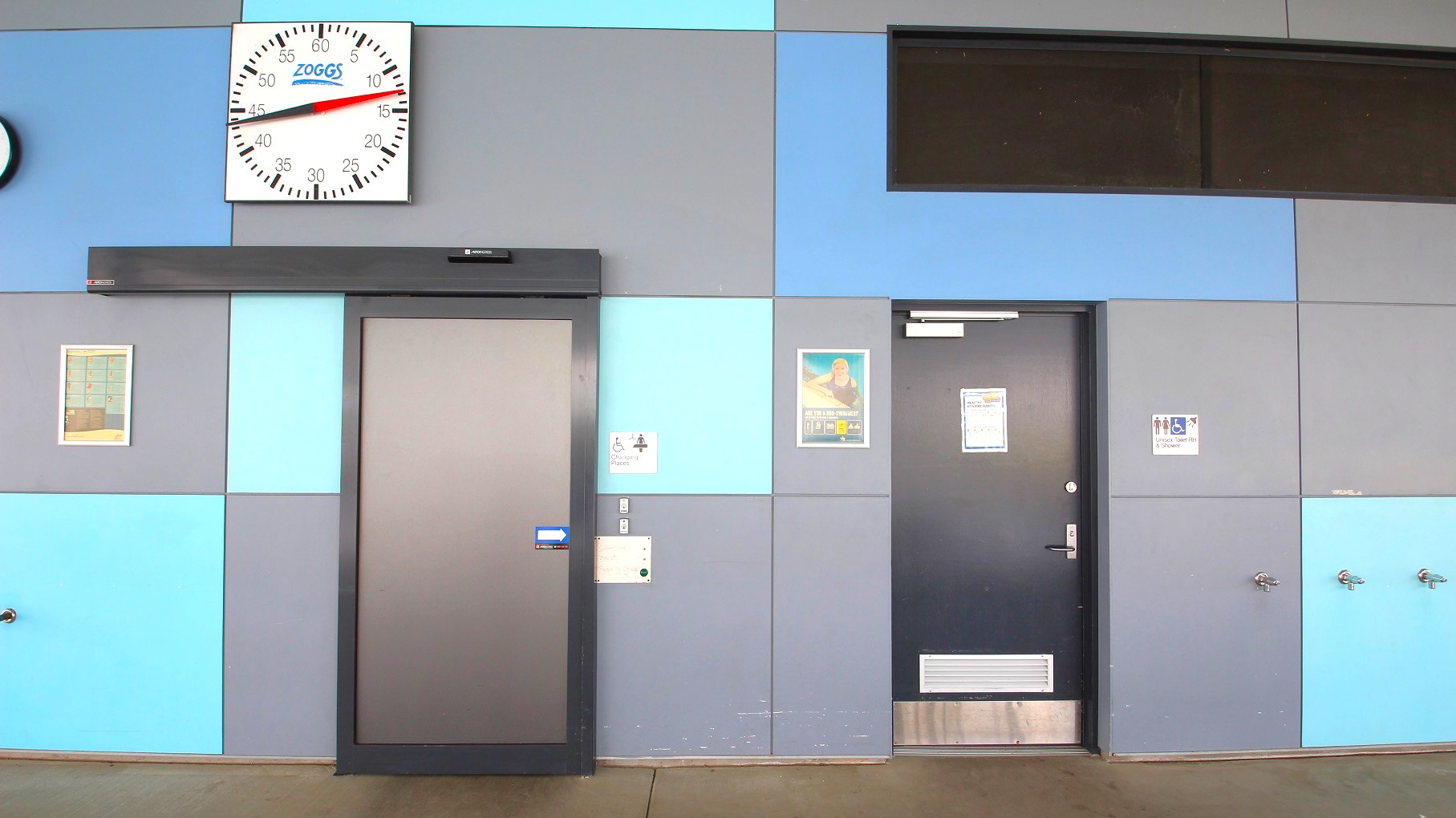 A wall with a geometric pattern of grey and blue panels. On the left, a large clock with "Zoggs" branding is mounted above a grey door. To the right, another door with a poster and a sign indicating accessible changing facilities. Further right, a sign for accessible toilets is visible. The floor is a plain brown surface, and there are small fixtures, possibly taps, on the lower right side of the wall. A wall with a geometric pattern of grey and blue panels. On the left, a large clock with "Zoggs" branding is mounted above a grey door. To the right, another door with a poster and a sign indicating accessible changing facilities. Further right, a sign for accessible toilets is visible. The floor is a plain brown surface, and there are small fixtures, possibly taps, on the lower right side of the wall.