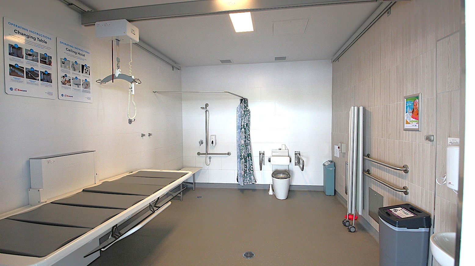 A spacious, accessible bathroom with a grey floor and white walls. On the left, there is a large changing table with padded sections and instructional posters above. In the centre, a ceiling hoist system is installed, and a shower area with a curtain is visible. On the right, there is a toilet with support rails, a bin, and folded equipment against the wall. The ceiling has a rectangular light fixture. A spacious, accessible bathroom with a grey floor and white walls. On the left, there is a large changing table with padded sections and instructional posters above. In the centre, a ceiling hoist system is installed, and a shower area with a curtain is visible. On the right, there is a toilet with support rails, a bin, and folded equipment against the wall. The ceiling has a rectangular light fixture.