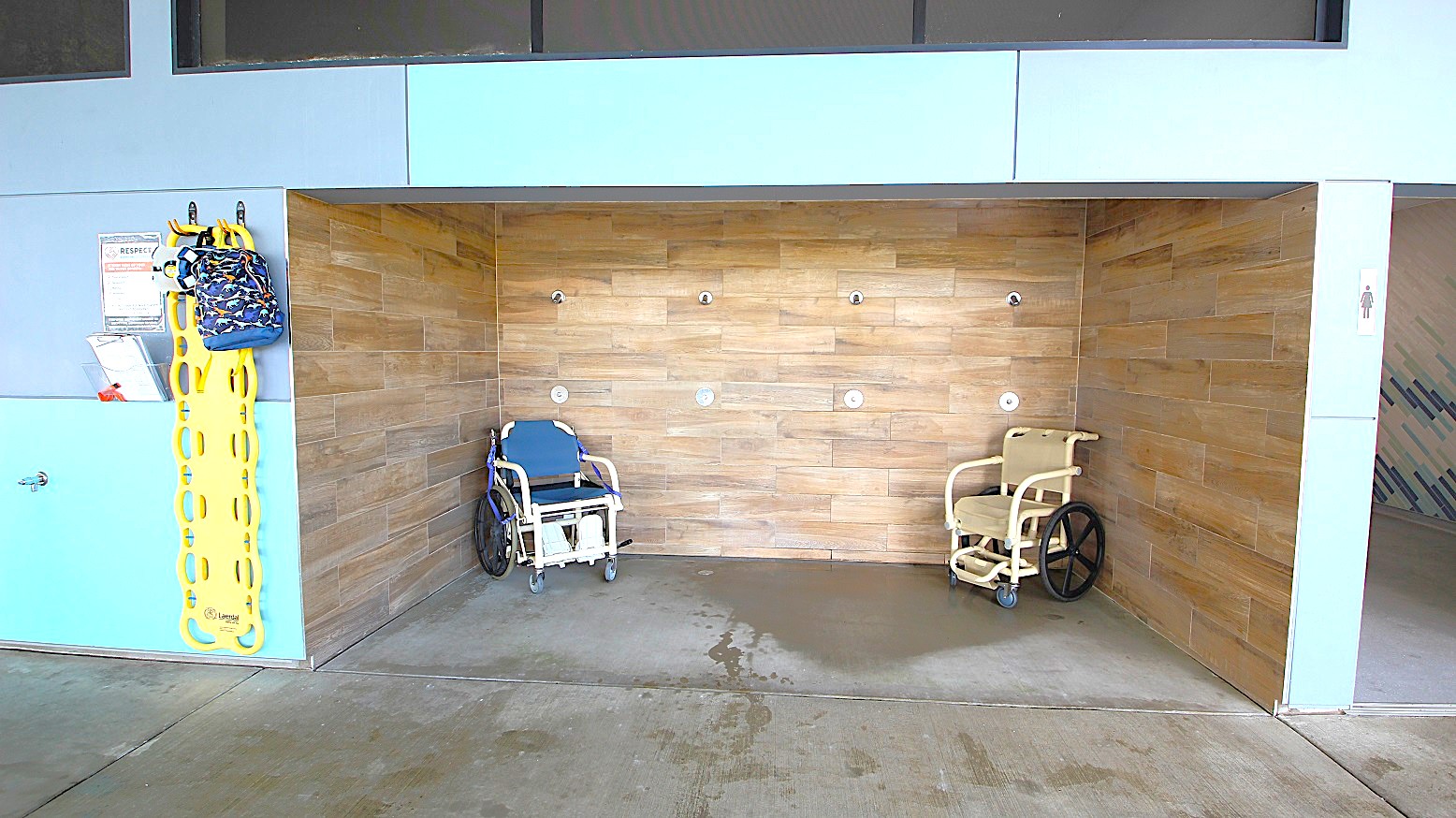 On the left, a yellow spine board and a colourful bag hang on a grey and blue wall, with a sign and a small shelf. In the middle, a wooden-tiled shower area with two shower chairs, one blue and one beige, is visible. The floor is concrete with water stains. The ceiling is not visible. On the left, a yellow spine board and a colourful bag hang on a grey and blue wall, with a sign and a small shelf. In the middle, a wooden-tiled shower area with two shower chairs, one blue and one beige, is visible. The floor is concrete with water stains. The ceiling is not visible.