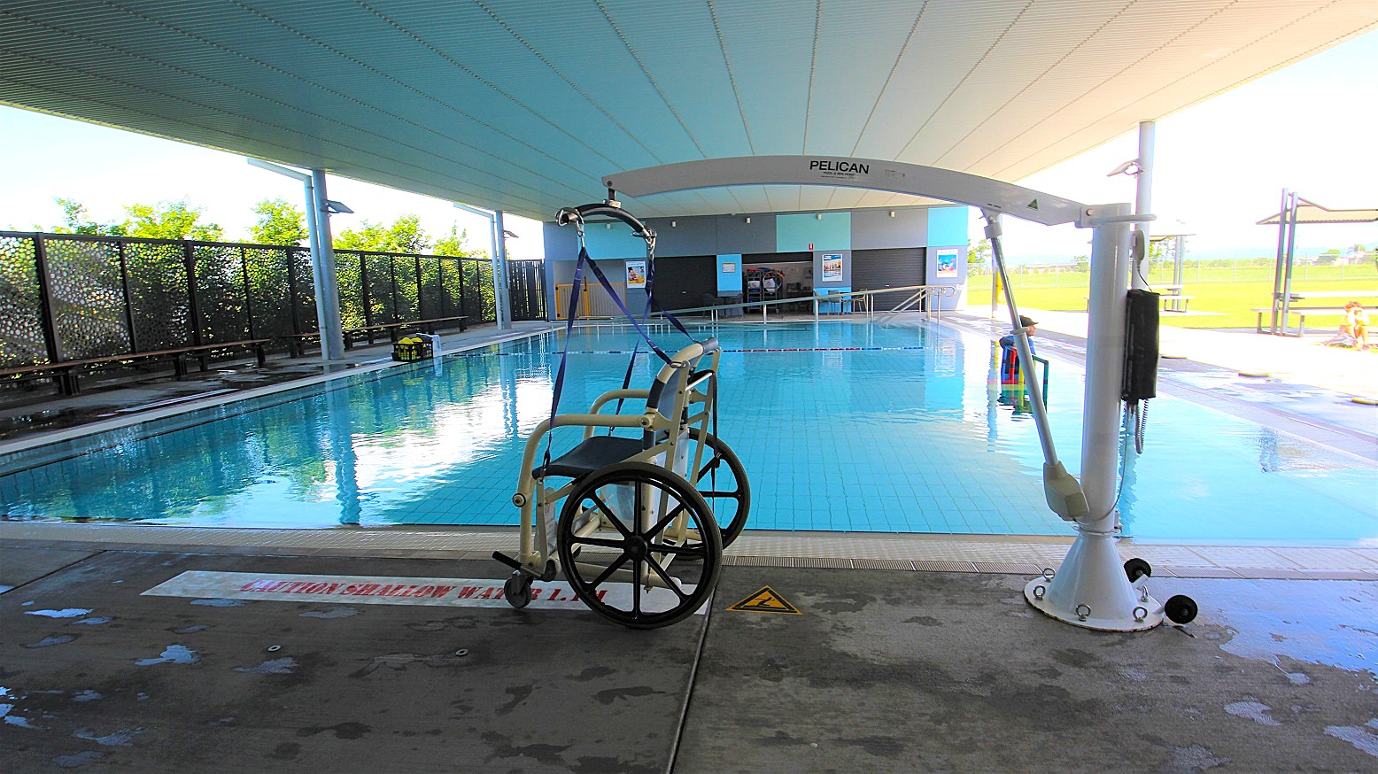 A swimming pool with a hoist system on the right, featuring a wheelchair attached to it. The pool is indoors with a high ceiling and large windows on the left, allowing natural light to enter. The floor is wet and has a caution sign. In the background, there are benches and a fence with greenery visible through it. The pool area is spacious and well-lit. A swimming pool with a hoist system on the right, featuring a wheelchair attached to it. The pool is indoors with a high ceiling and large windows on the left, allowing natural light to enter. The floor is wet and has a caution sign. In the background, there are benches and a fence with greenery visible through it. The pool area is spacious and well-lit.
