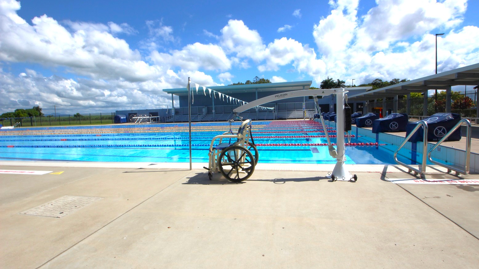 A swimming pool with multiple lanes is in the centre, with lane dividers visible. In the foreground, a wheelchair lift is positioned on the pool deck, next to a wheelchair. On the right, there are steps leading into the pool. The background features a building with a shaded area and a row of flags strung above the pool. The ground is a light-coloured concrete surface. A swimming pool with multiple lanes is in the centre, with lane dividers visible. In the foreground, a wheelchair lift is positioned on the pool deck, next to a wheelchair. On the right, there are steps leading into the pool. The background features a building with a shaded area and a row of flags strung above the pool. The ground is a light-coloured concrete surface.