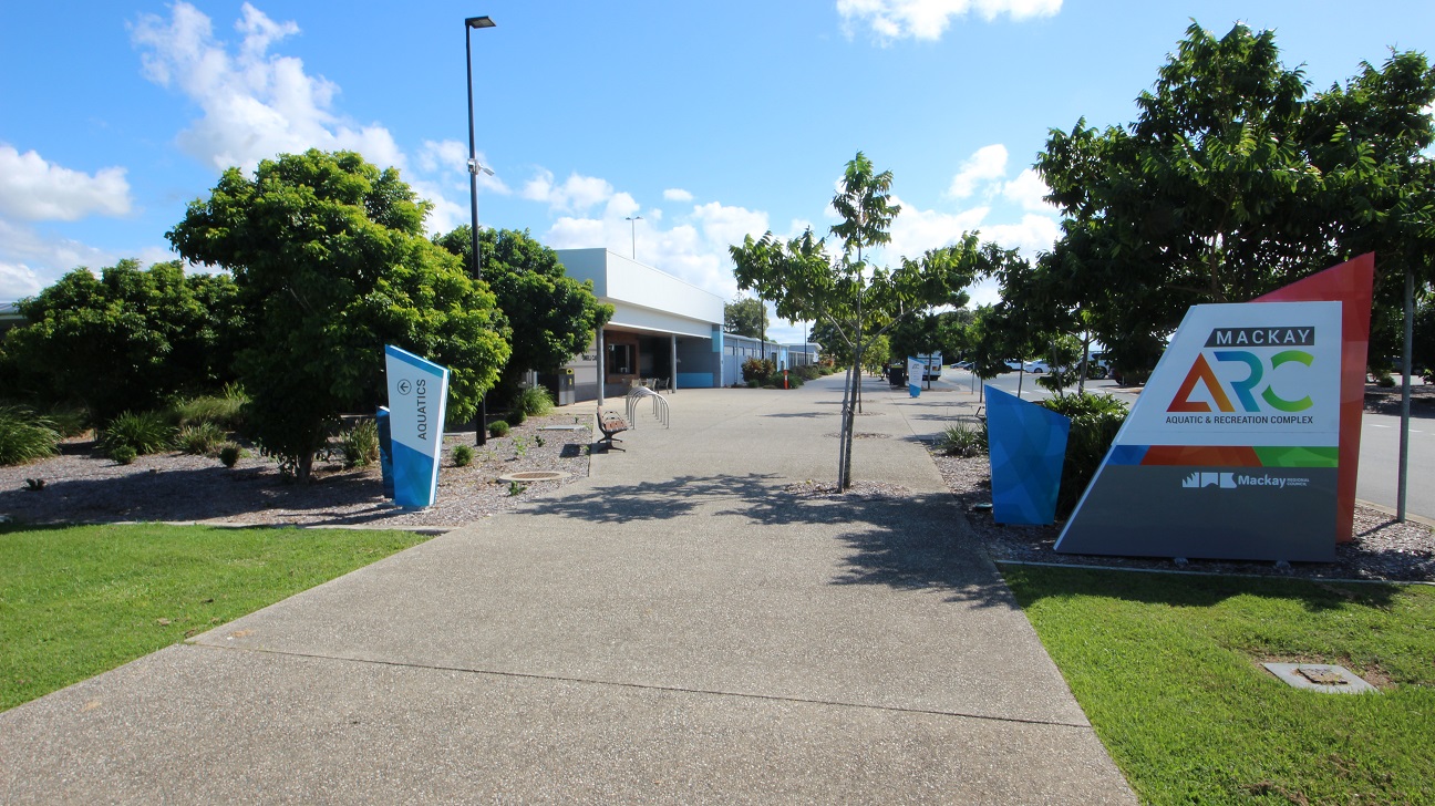 A straight concrete road flanked by green grass and trees. On the right, a concrete structure displays facility signage. Additional concrete signages, including one directing to Aquatics, are adjacent. Trees, street lights, and benches line the left side. Further ahead, a low-set building comes into view. A straight concrete road flanked by green grass and trees. On the right, a concrete structure displays facility signage. Additional concrete signages, including one directing to Aquatics, are adjacent. Trees, street lights, and benches line the left side. Further ahead, a low-set building comes into view.