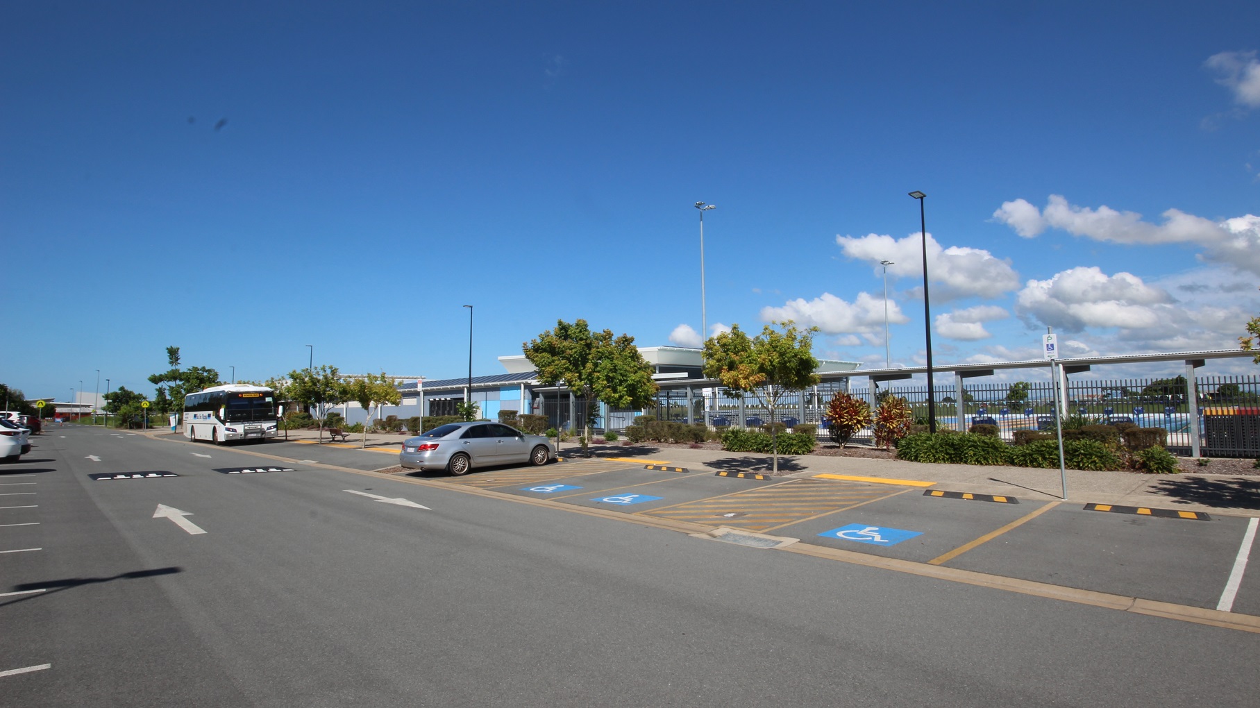 A road with parking on both sides. To the left, cars are parked, occupying several spaces. On the right, accessible parking slots with blue wheelchair signs and yellow diagonal lines are visible. A bus and a grey vehicle are currently parked. Trees and a fence border the right side, and behind is an aquatic centre. Tall street lights are visible. A road with parking on both sides. To the left, cars are parked, occupying several spaces. On the right, accessible parking slots with blue wheelchair signs and yellow diagonal lines are visible. A bus and a grey vehicle are currently parked. Trees and a fence border the right side, and behind is an aquatic centre. Tall street lights are visible.