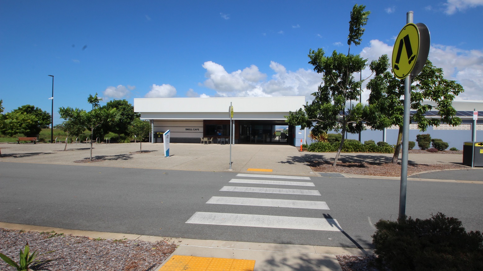 A concrete road with pedestrian markings leads to a low-rise building with a white roof, featuring a step-free ramp. Yellow truncated domes mark both ends of the lane. Road signs line the road edge. Bike racks, a bench chair, and tall street lights are visible on the far left. Trees and trimmed shrubs are on the right. A concrete road with pedestrian markings leads to a low-rise building with a white roof, featuring a step-free ramp. Yellow truncated domes mark both ends of the lane. Road signs line the road edge. Bike racks, a bench chair, and tall street lights are visible on the far left. Trees and trimmed shrubs are on the right.