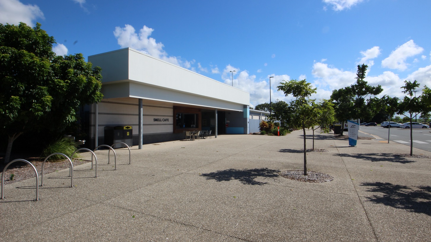 On the left is a low-rise facility building with an extended white roof. Tables, chairs, and rubbish bins are under the shade, along with bike racks positioned in front of a tree and grass. To the right, vehicles parked in the car park are visible. Trees line the front and sides, with a retractable banner in place. On the left is a low-rise facility building with an extended white roof. Tables, chairs, and rubbish bins are under the shade, along with bike racks positioned in front of a tree and grass. To the right, vehicles parked in the car park are visible. Trees line the front and sides, with a retractable banner in place.