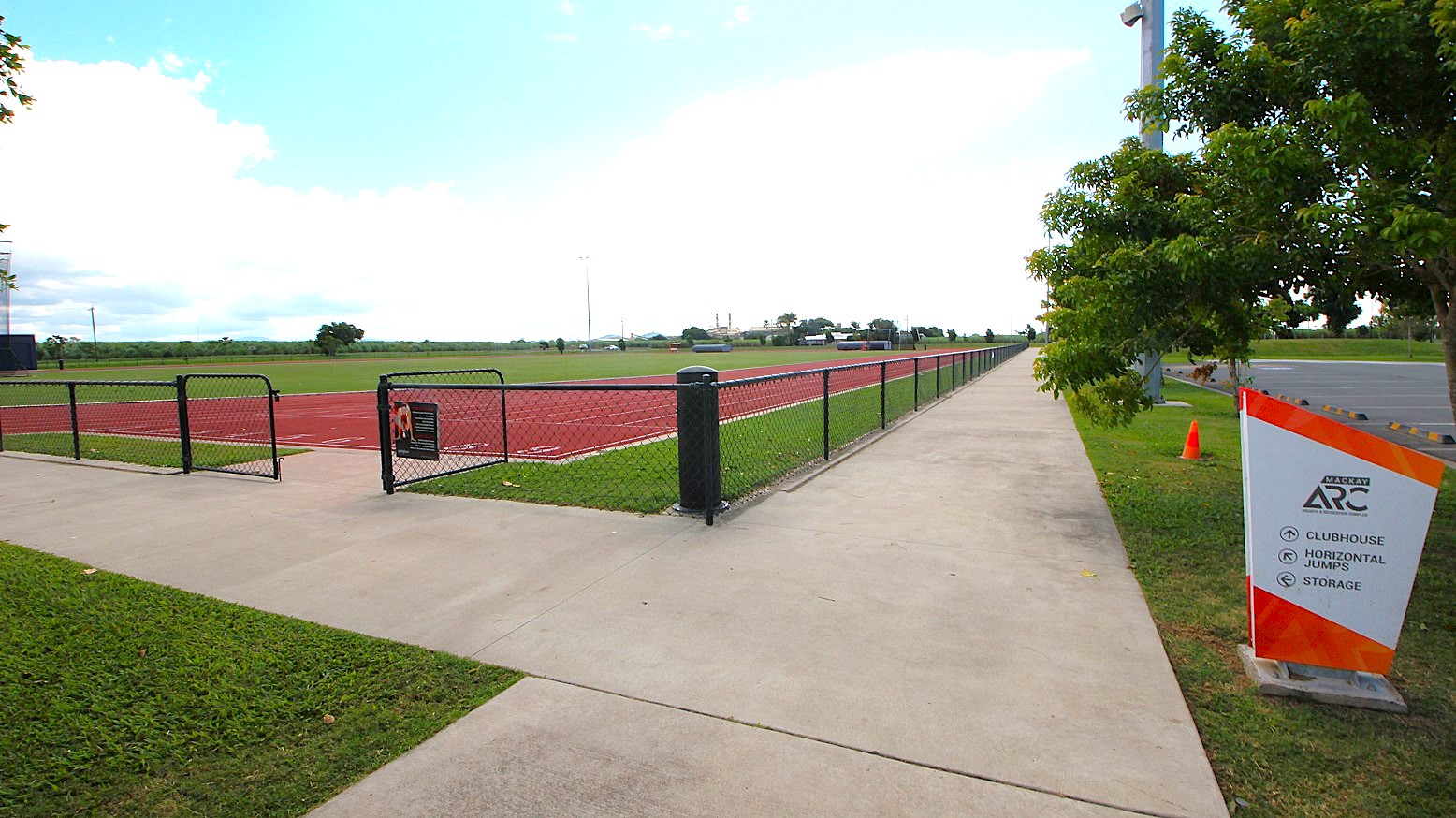 A paved walkway runs alongside a fenced athletic track on the left, with a grassy field inside. On the right, a sign with directional information stands near a row of trees. The path extends into the distance, bordered by a parking area. A single orange cone is visible near the sign. A paved walkway runs alongside a fenced athletic track on the left, with a grassy field inside. On the right, a sign with directional information stands near a row of trees. The path extends into the distance, bordered by a parking area. A single orange cone is visible near the sign.