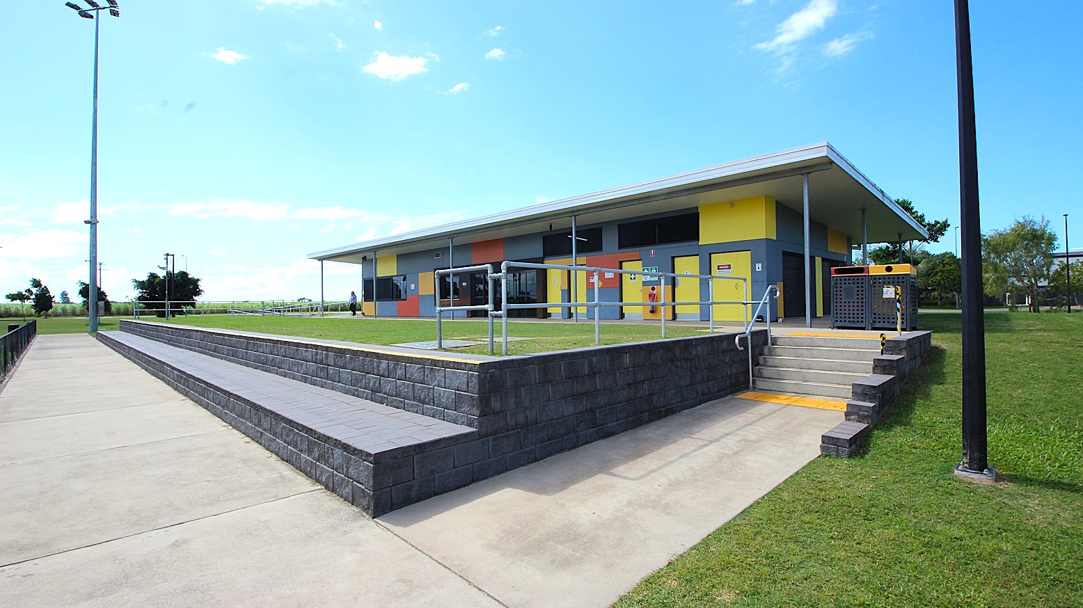A modern building with a colourful facade featuring yellow, orange, and blue panels. In the foreground, there is a concrete pathway and a low brick wall with steps leading up to the building. Metal railings are present near the steps. The surrounding area is grassy, with a tall streetlight on the left. The sky is clear with a few clouds. A modern building with a colourful facade featuring yellow, orange, and blue panels. In the foreground, there is a concrete pathway and a low brick wall with steps leading up to the building. Metal railings are present near the steps. The surrounding area is grassy, with a tall streetlight on the left. The sky is clear with a few clouds.